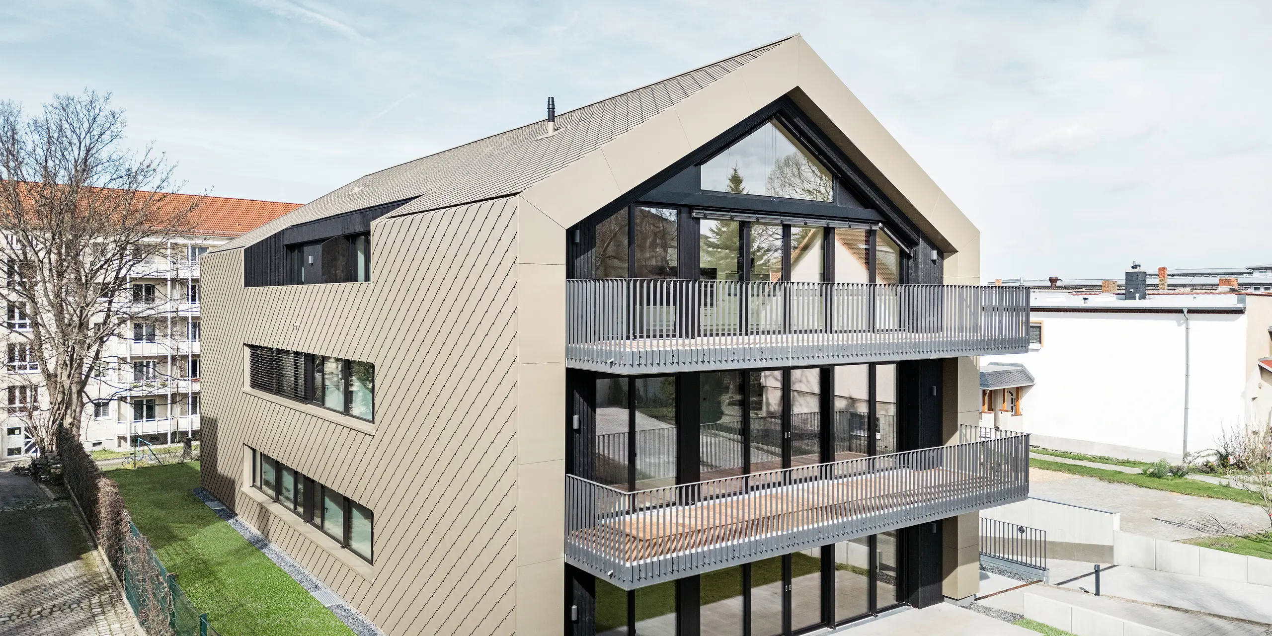 Pohled do zahrady na moderní bytový dům na Chemnitzer Straße v Drážďanech. Udržovaná zahrada je obklopena působivou architekturou. Fasáda a střecha budovy jsou opláštěny robustní falcovanou a fasádní šablonou 29 × 29 v bronzové barvě. Hliníkové šablony PREFA dodávají konstrukci elegantní vzhled. Obrázek zobrazuje prostorné balkony, velká okna a harmonické začlenění budovy do okolního prostoru.