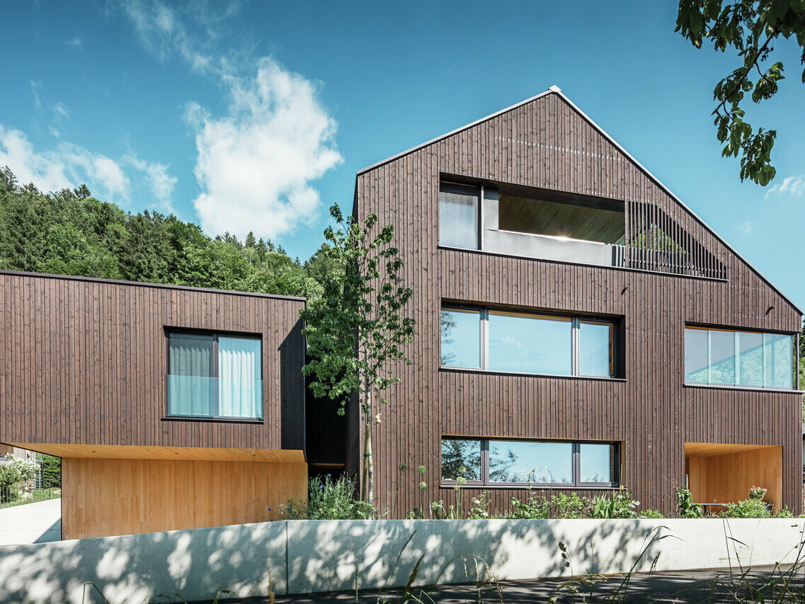 Moderní bytový dům s ořechově hnědou střechou PREFA a tmavou dřevěnou fasádou, proložený světlými dřevěnými prvky, velkými okny a obklopený zelení.