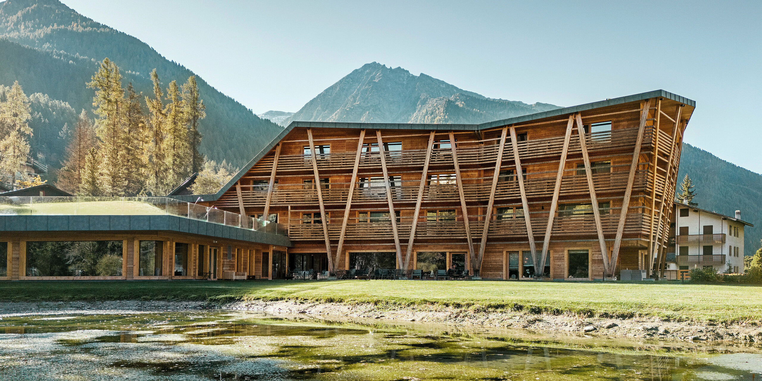 Das Bild zeigt die beeindruckende Pracht des 'Au Charmant Petit Lac', eines Luxus-Öko-Hotels in Champoluc, Italien, vor dem Hintergrund majestätischer Berge. Das Hotel zeichnet sich durch eine ausdrucksstarke Holzarchitektur aus, die sich nahtlos in die natürliche Umgebung einfügt. Der kleine See im Vordergrund spiegelt die ruhige und entspannende Atmosphäre wider, die das Hotel umgibt. Die umweltbewusste Bauweise wird durch die Nutzung von PREFALZ in P.10 Steingrau für das Dach unterstrichen, was die nachhaltige Philosophie des 5-Sterne-Hauses betont.