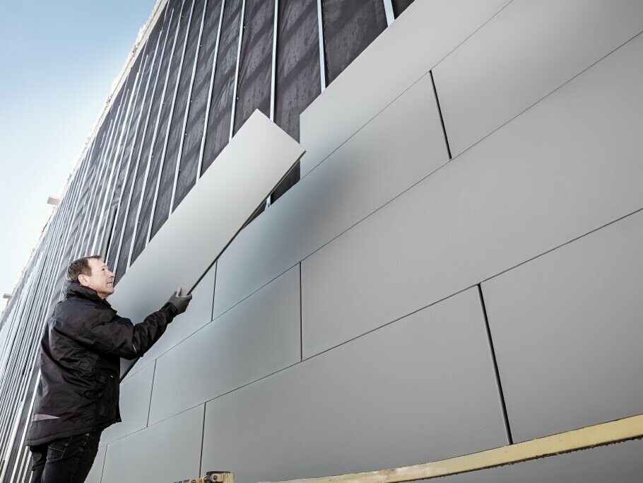 Fotografie částečně hotové fasády obložené lamelami PREFA Siding v šířce 500. Klempíř pokládá další fasádní lamelu, snímek je pořízen zespodu.