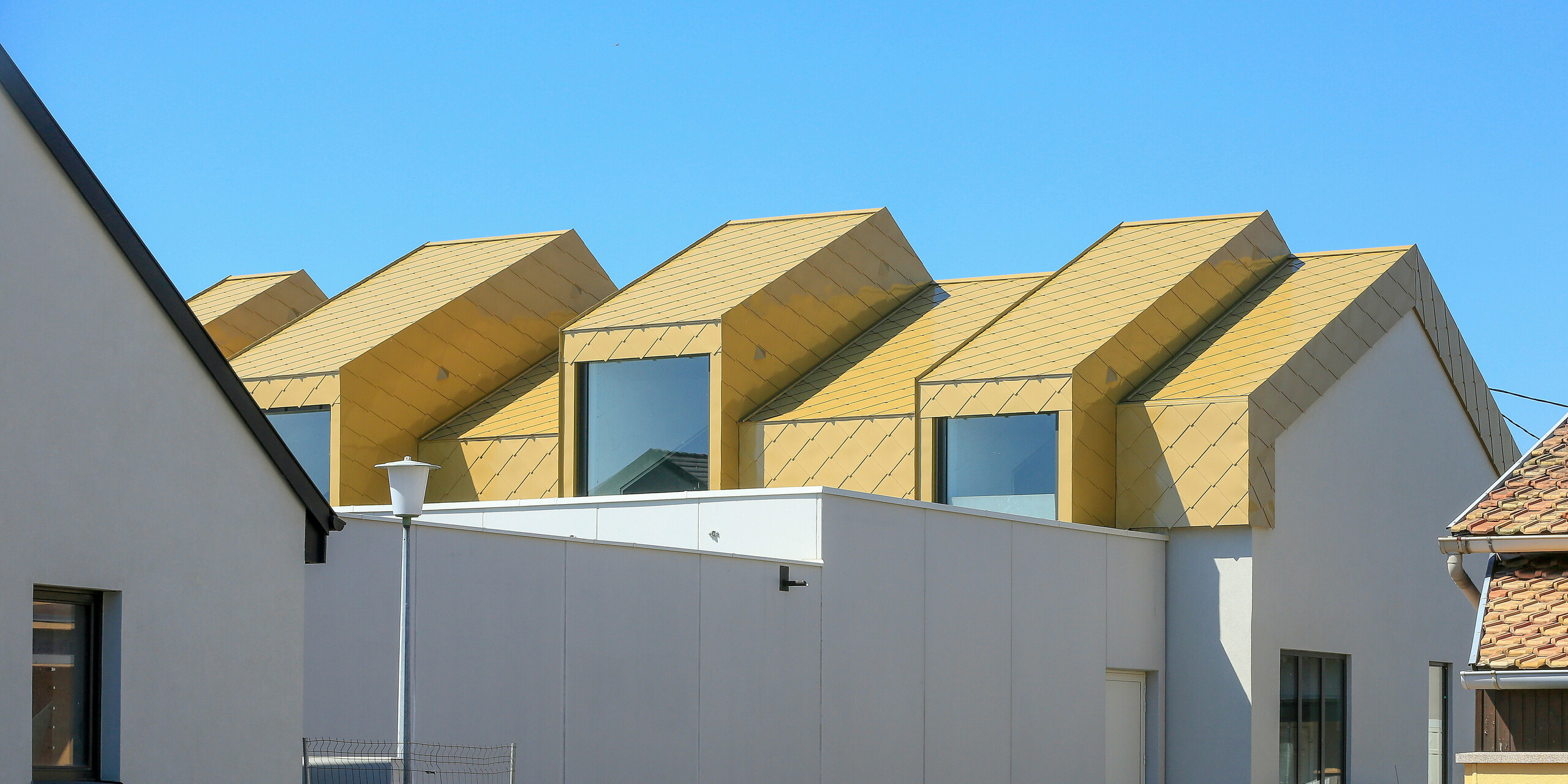 Pohled z dálky na budovu s lesklou střechou PREFA a fasádními šablonami 44 × 44 v provedení Sparkling Gold (barvě na přání)