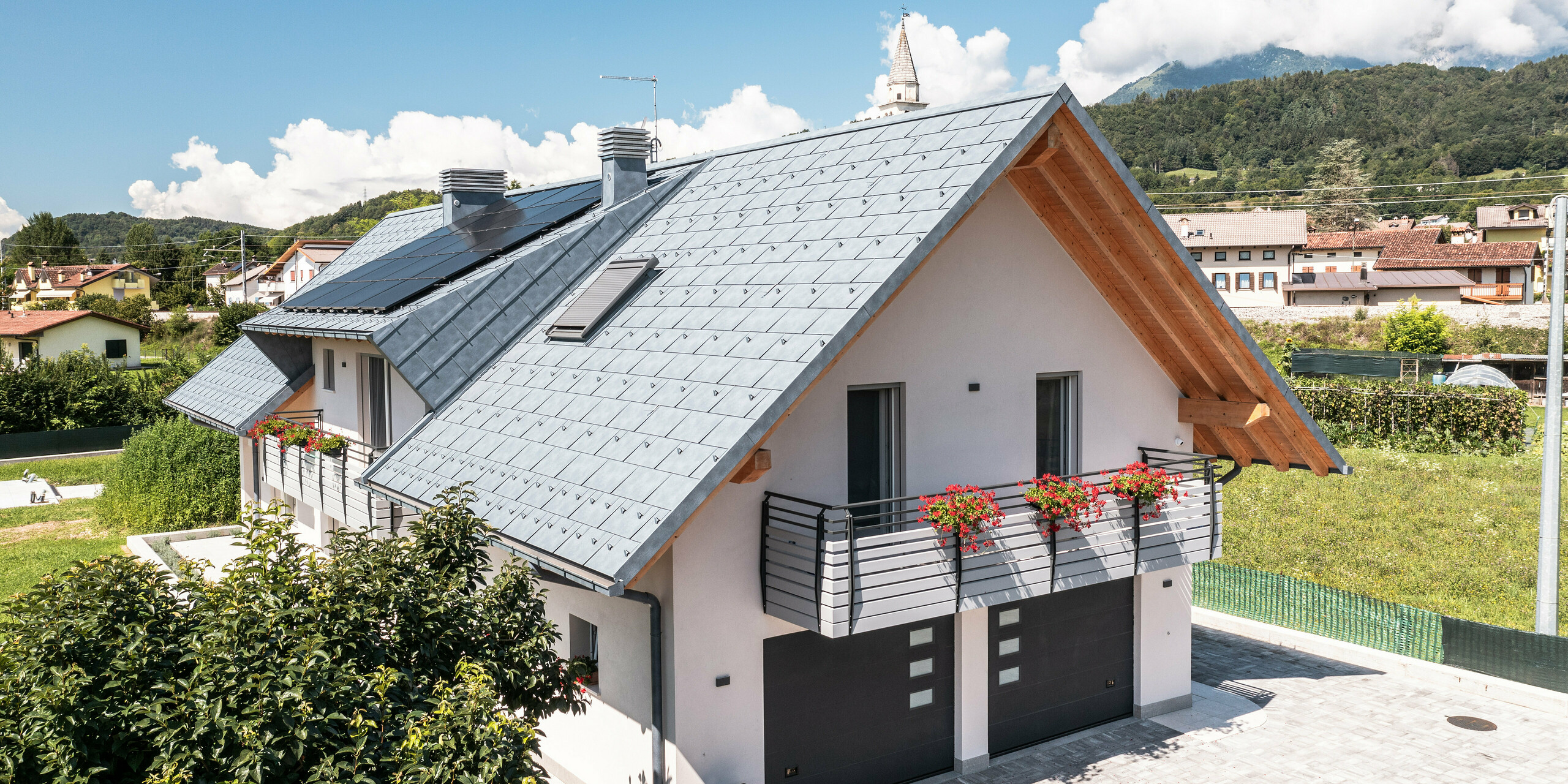 Moderní rodinný dům v italském Bellunu, charakteristický střešním systémem PREFA v antracitové barvě v podobě střešního panelu R.16, který opticky ladí s viditelnými dřevěnými trámy štítu. Pečlivě navržený venkovní prostor s kvetoucími balkónovými truhlíky a dobře udržovaným živým plotem dodává nemovitosti lákavý šmrnc, zasazený do klidné vesnice na pozadí Dolomit. Pod střechou jsou dva vjezdy do garáže. Barva garážových vrat ladí s hliníkovými prvky a dodává nemovitosti celkový harmonický dojem.