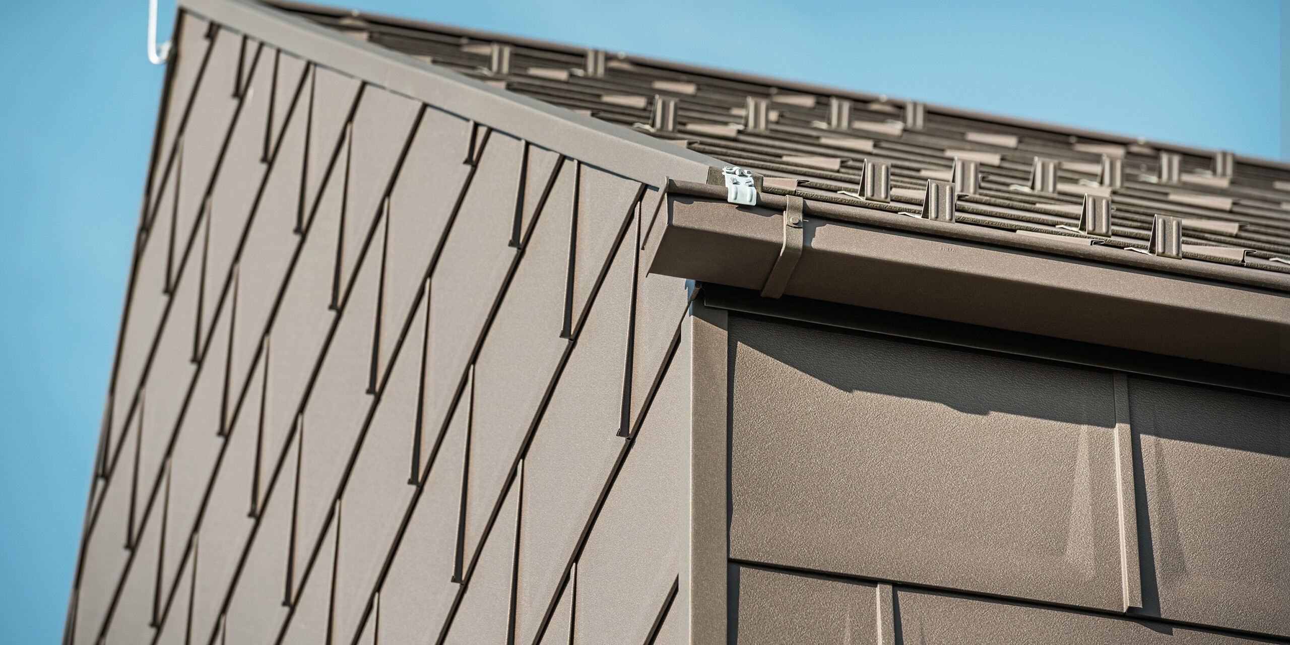 Detailní pohled na odvodnění střechy rodinného domu v rakouském Neukirchenu se schránkovým žlabem PREFA. Střešní panel R.16 v hnědé barvě P.10 tvoří nejen střechu, ale také zdobí fasádu a vytváří konzistentní a elegantní vzhled. Tento detailní záběr ukazuje preciznost a kvalitu hliníkových výrobků PREFA, které spojují design s funkčností pro moderní pojetí bydlení.