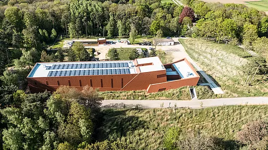 Letecký pohled na čerpací stanici pitné vody WML v Lottum, pozoruhodný infrastrukturní prvek harmonicky integrovaný do zelené krajiny lesů a luk. Protáhlá budova s ​​charakteristickou hliníkovou fasádou PREFA Siding.X v teplé, rezavě zbarvené povrchové úpravě působí jako sochařský orientační bod. Mezi poutavé prvky patří jasně strukturovaný tvar budovy, integrované fotovoltaické moduly na střeše a jemně fazetovaný povrch fasády. Odolné, lehké a recyklovatelné hliníkové desky PREFABOND nabízejí výjimečnou odolnost vůči povětrnostním vlivům, udržitelnou účinnost a nenáročný na údržbu plášť budovy.