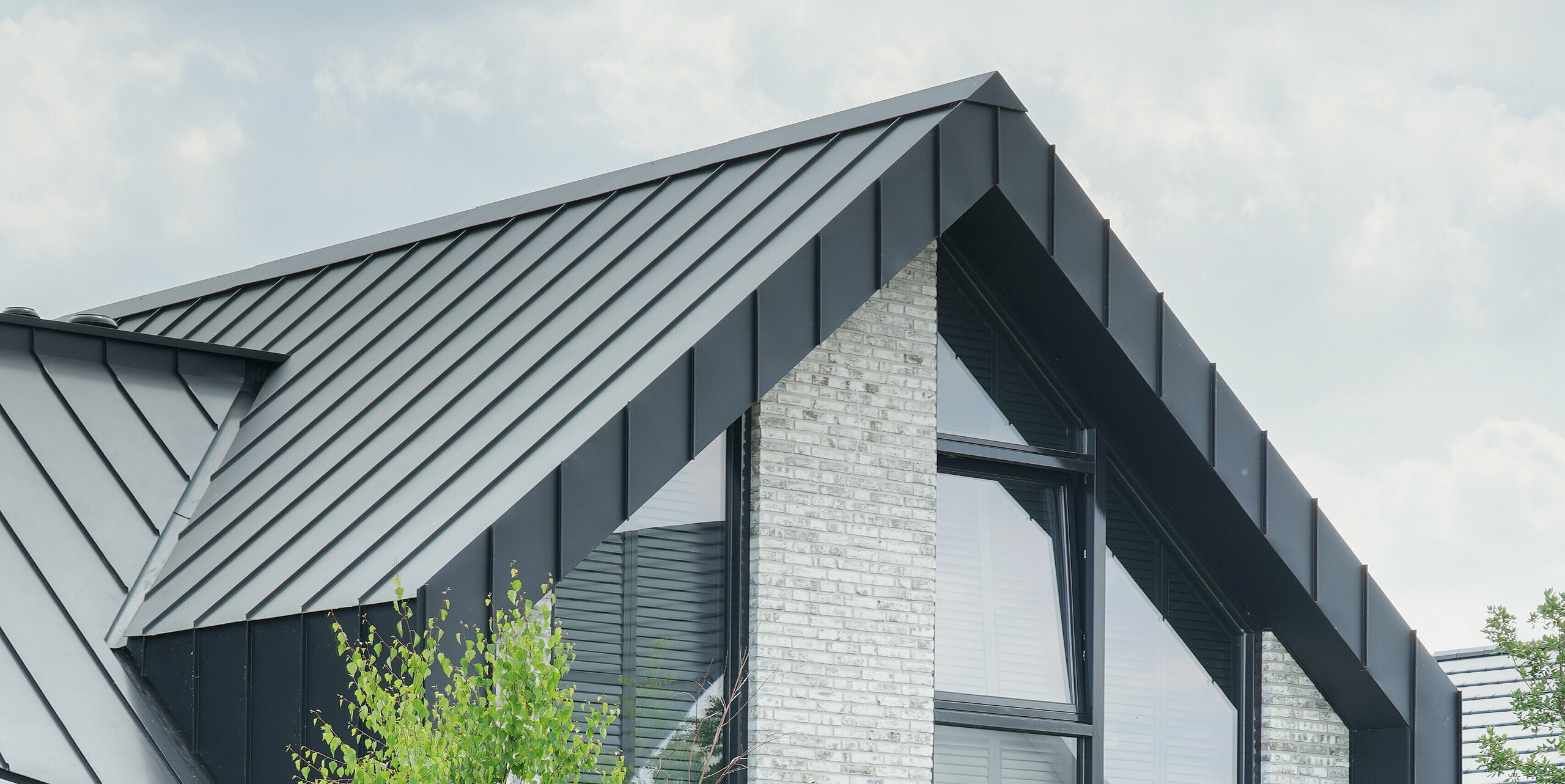 Částečný pohled na moderní obytnou budovu ve Veenendaal, Nizozemsko, s černým pláštěm budovy PREFALZ. Kombinace ostře zkosených linií střechy a strukturované, světle šedé fasády z přírodního kamene vytváří vzrušující architektonickou hru. Velká okna od podlahy ke stropu prolamují fasádu a umožňují výhled do přírody.