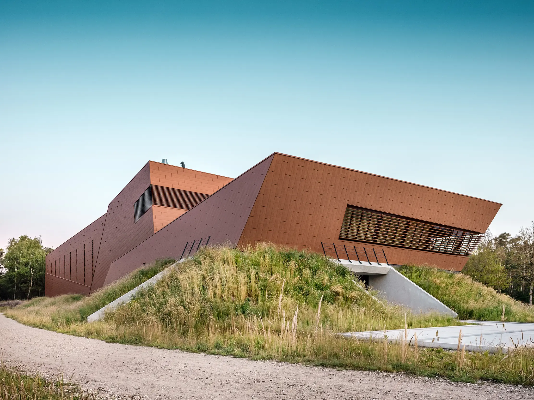 Architektonicky expresivní čerpací stanice pitné vody WML v Lottum se vyznačuje sochařsky skládanými tvary budov, zasazenými do přírodní krajiny nábřeží. Výrazná hliníková fasáda PREFA Siding.X s teplým, rezavějícím vzhledem a fazetovaným, zářivým povrchem je skutečným poutačem a vytváří působivou hru světla a stínu. Horizontální žaluzie a přesné linie zdůrazňují dynamickou geometrii. Lehké, trvanlivé a plně recyklovatelné hliníkové panely PREFA nabízejí maximální odolnost proti povětrnostním vlivům, udržitelnou konstrukci a nenáročný na údržbu pláště budovy.