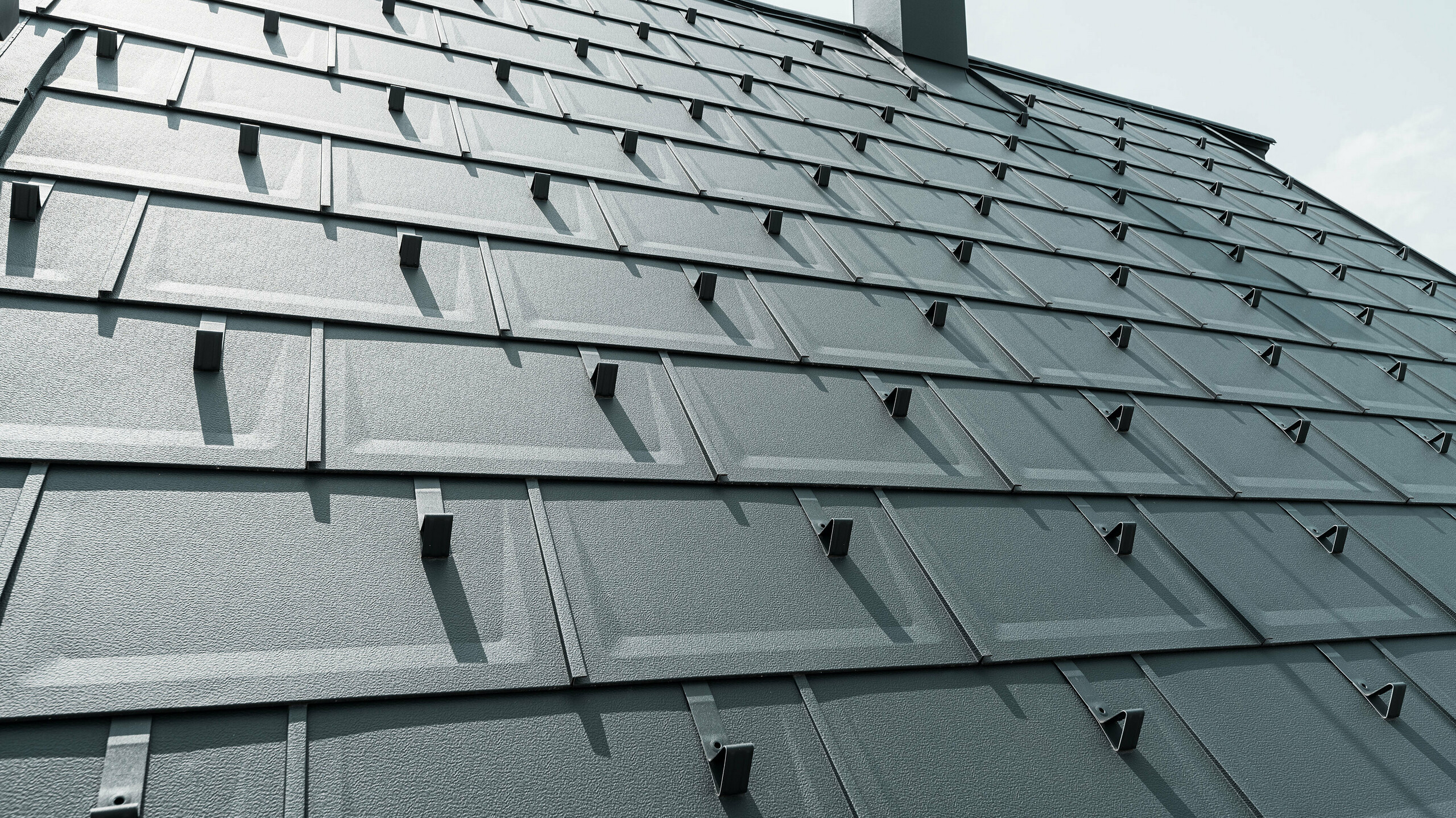 Detailní záběr střešní panelu PREFA R.16 v barvě P.10 antracitová. Na střeše jsou vidět sněhové zábrany PREFA a komínová krytina.