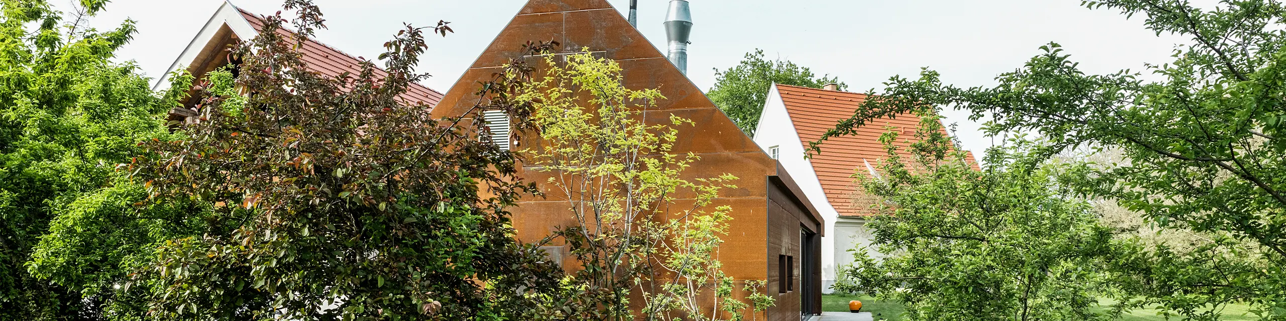 Rezidence Velich v Seewinkel, Burgenland: Pohled do moderních zahrad s krásnými rostlinami. Rezidence slouží jako místo pro odborný dialog PREFARENZEN 2025, kde architekti a odborníci PREFA vybírají ty nejzajímavější architektonické objekty. V popředí můžete vidět stylovou, rezavě zbarvenou kovovou stěnou s napsáním 'RESIDENCE VELICH'. Architektura budovy kombinuje tradiční a moderní prvky a harmonicky zapadá do přírodního prostředí.