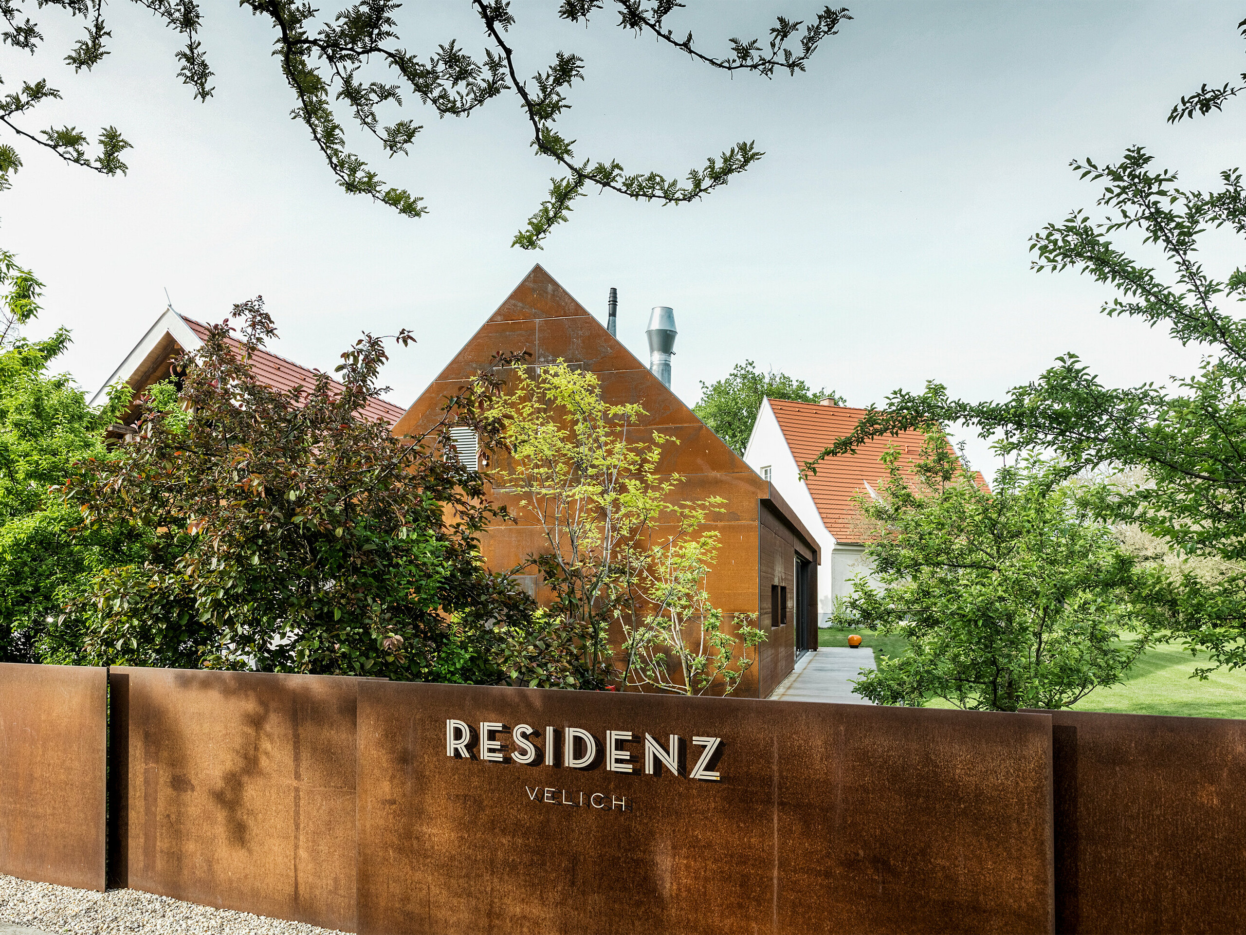 Rezidence Velich v Seewinkel, Burgenland: Pohled do moderních zahrad s krásnými rostlinami. Rezidence slouží jako místo pro odborný dialog PREFARENZEN 2025, kde architekti a odborníci PREFA vybírají ty nejzajímavější architektonické objekty. V popředí můžete vidět stylovou, rezavě zbarvenou kovovou stěnou s napsáním 'RESIDENCE VELICH'. Architektura budovy kombinuje tradiční a moderní prvky a harmonicky zapadá do přírodního prostředí.