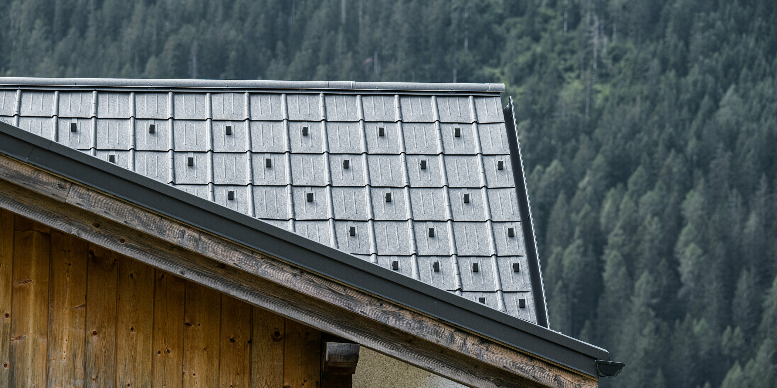 Detailní záběr hliníkové střechy se světle šedými falcovanými taškami od PREFA na horské chatě v Auronzo di Cadore v Itálii, obklopené hustými lesy. Precizní montáž a jednoduchý design střešních panelů zdůrazňuje venkovskou architekturu a zároveň poskytuje moderní ochranu a estetiku v souladu s přírodním horským prostředím.