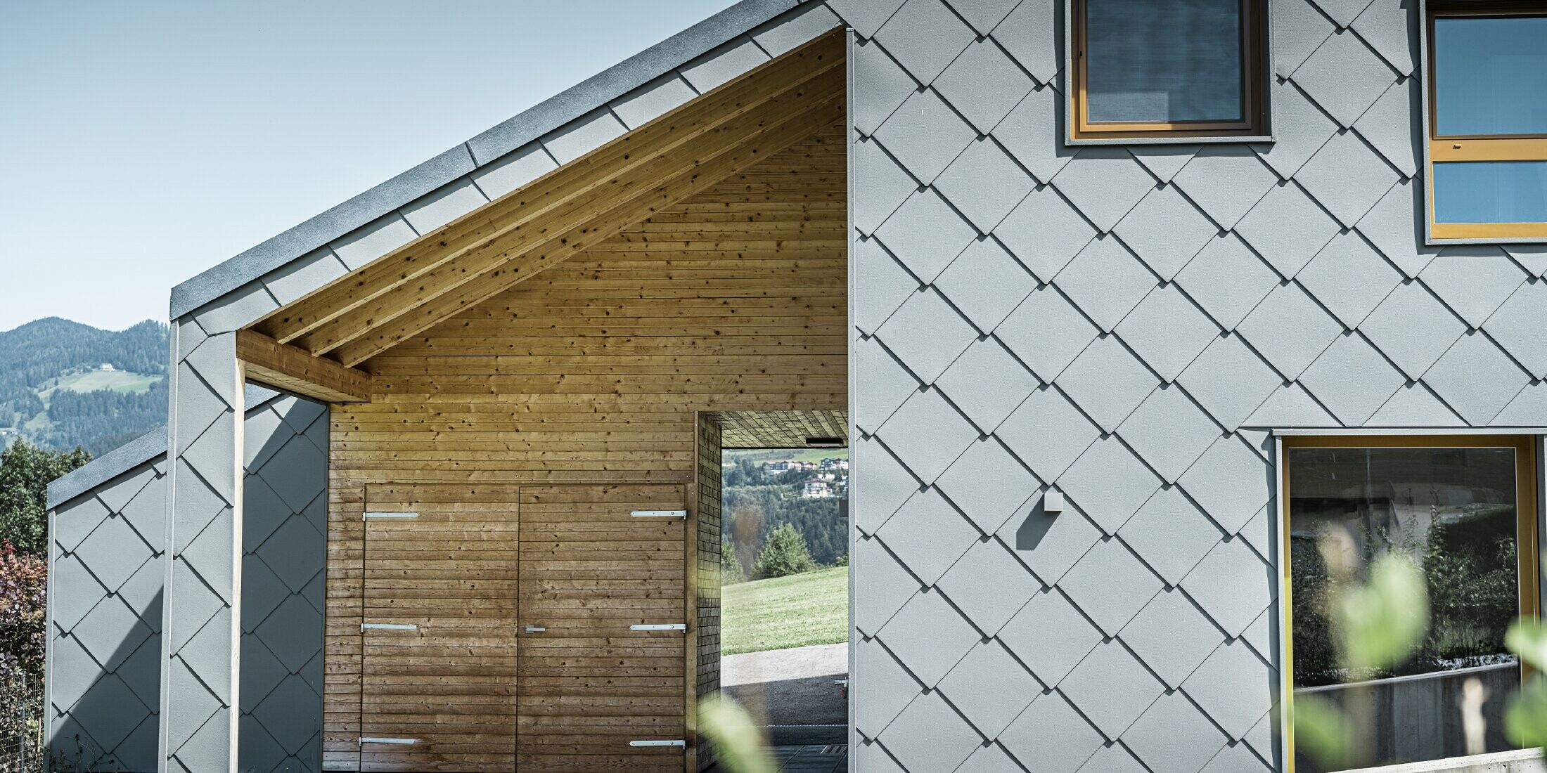 Zakrytý vstupní prostor je proveden horizontálním dřevěným obkladem, zbytek fasády je opláštěn velkformátovou hliníkovou fasádní šablonou 44 x 44 PREFA ve světle šedé barvě