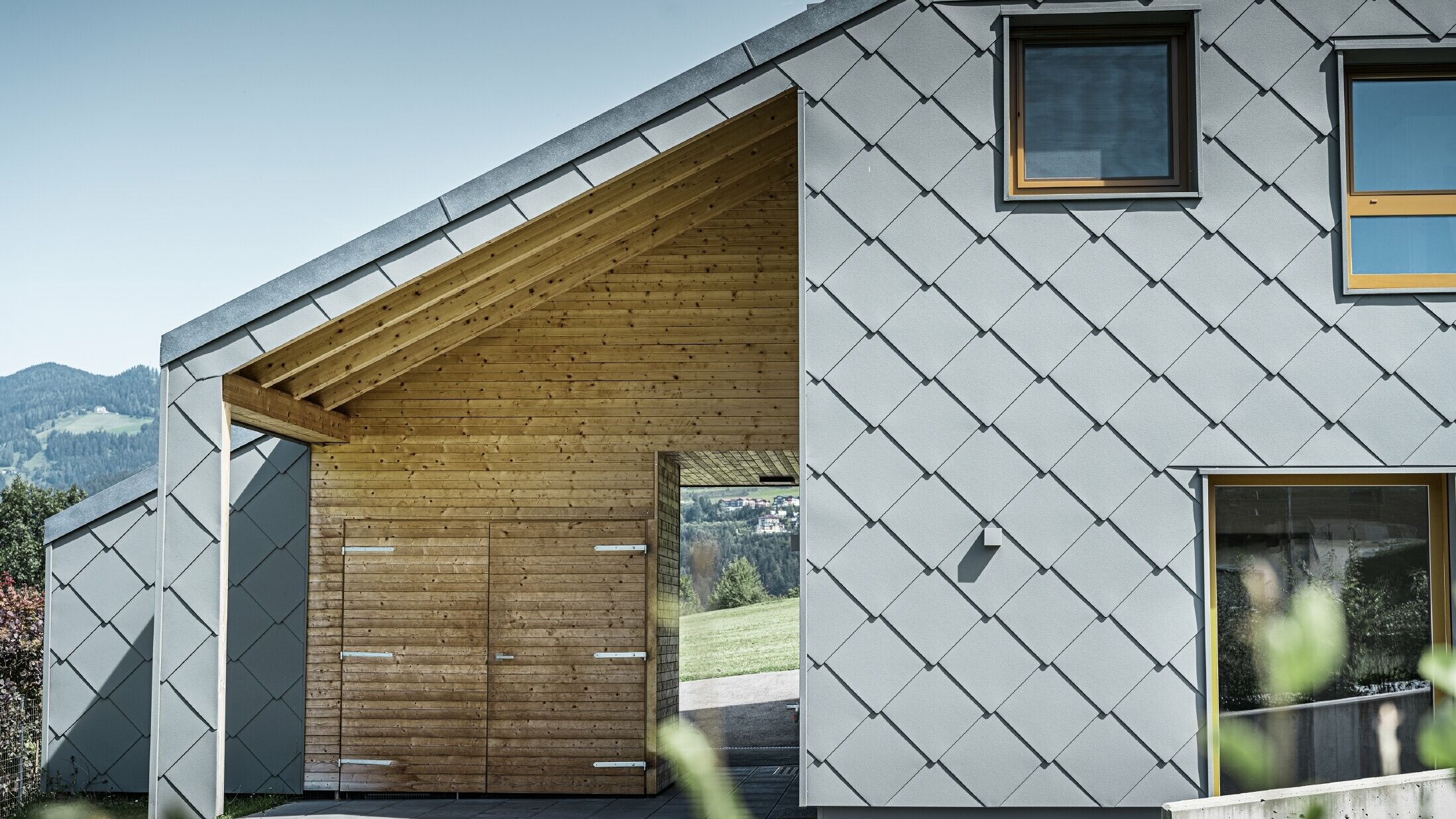 Zakrytý vstupní prostor je proveden horizontálním dřevěným obkladem, zbytek fasády je opláštěn velkformátovou hliníkovou fasádní šablonou 44 x 44 PREFA ve světle šedé barvě