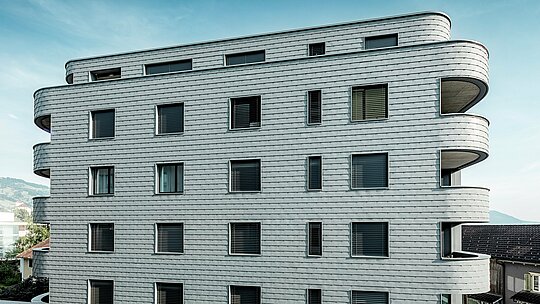 Čelní pohled na Bernerhaus v Buchsu s jemně zaoblenými rohy budovy a souvislou fasádou z PREFA fasádních šindelů v barvě P.10 světle šedé. Šupinatá hliníková fasáda vytváří zářivý vzor a zdůrazňuje horizontální linie zakřivených balkonových pásů. Precizně integrované okenní otvory harmonicky splývají s homogenním povrchem. Díky lehkému, povětrnostním vlivům odolnému a trvanlivému hliníku PREFA vzniká nenáročný, rozměrově stabilní plášť budovy s výrazným vzhledem na alpském pozadí.