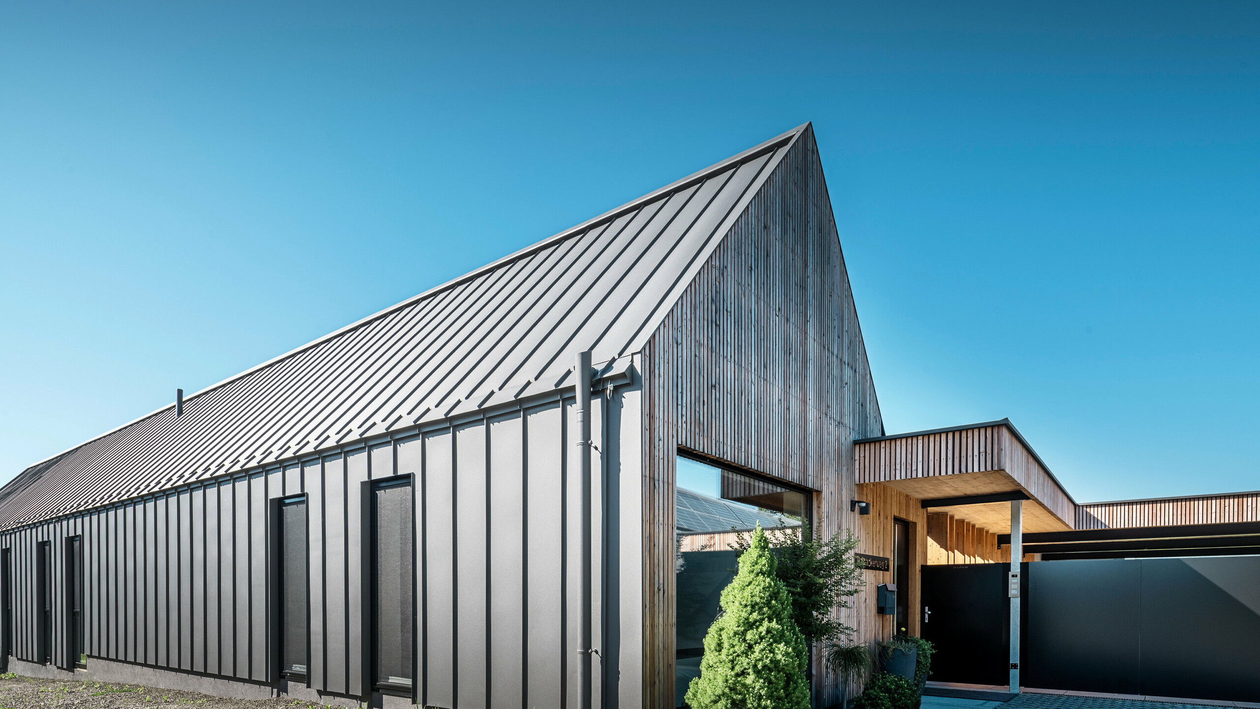 [Translate to Český:] Eingangsbereich eines Einfamilienhauses mit angrenzender Garage in Pogier, Österreich, inmitten einer malerischen Landschaft. Dach und Fassade beider Gebäude sind mit langlebigem PREFALZ in dunkelgrau P.10 eingedeckt, was ein modernes Erscheinungsbild ergibt. Die Garage hat eine harmonisch integrierte Holzverkleidung, die sich nahtlos in die umgebende Landschaft einfügt. Der gepflasterte Hof und die klaren Linien des Entwurfs runden die Ästhetik des Grundstücks ab, während die sanften Hügel im Hintergrund das Bild einer ruhigen grünen Oase im grünen Herzen Österreichs vervollständigen.