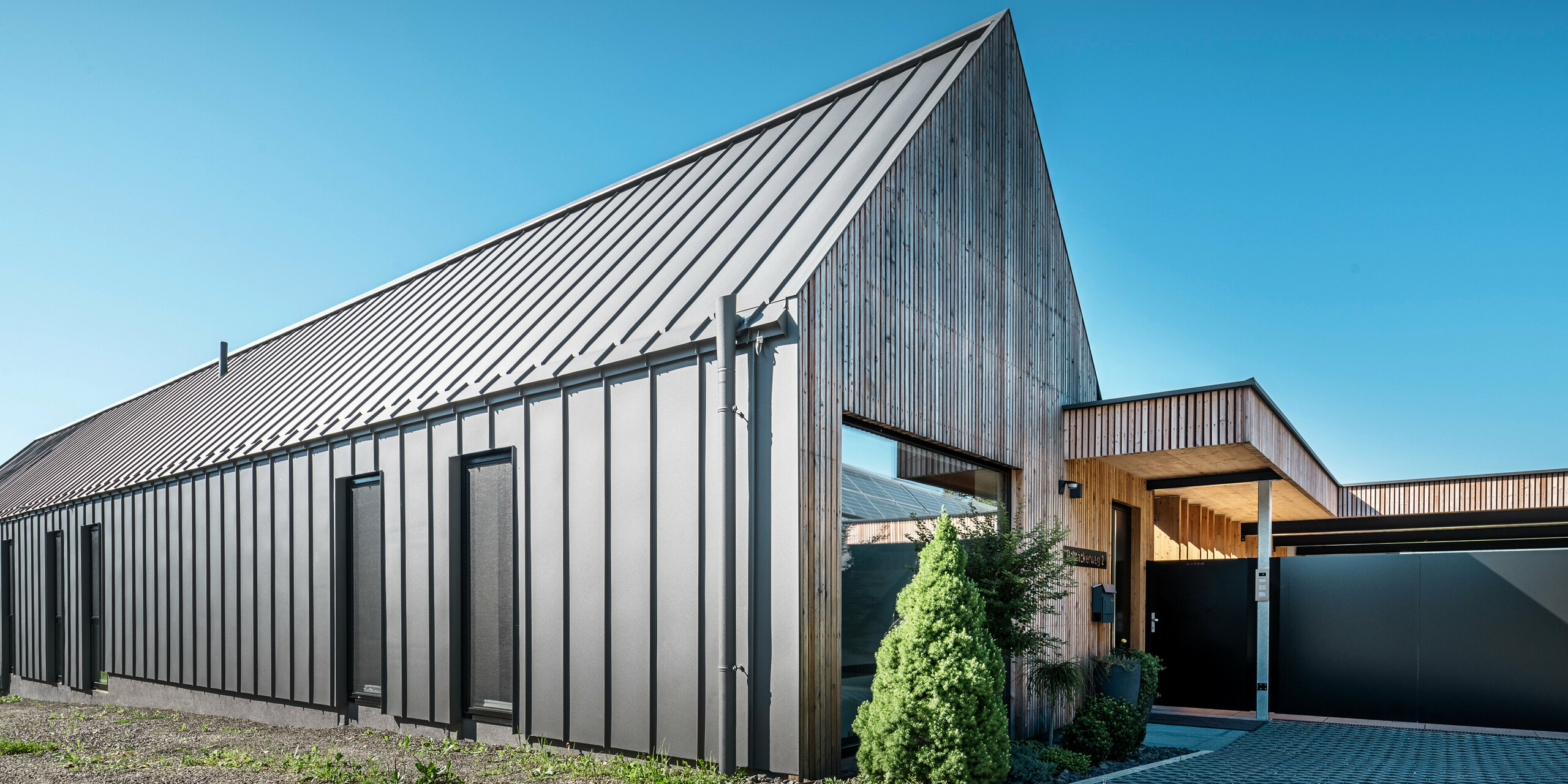 [Translate to Český:] Eingangsbereich eines Einfamilienhauses mit angrenzender Garage in Pogier, Österreich, inmitten einer malerischen Landschaft. Dach und Fassade beider Gebäude sind mit langlebigem PREFALZ in dunkelgrau P.10 eingedeckt, was ein modernes Erscheinungsbild ergibt. Die Garage hat eine harmonisch integrierte Holzverkleidung, die sich nahtlos in die umgebende Landschaft einfügt. Der gepflasterte Hof und die klaren Linien des Entwurfs runden die Ästhetik des Grundstücks ab, während die sanften Hügel im Hintergrund das Bild einer ruhigen grünen Oase im grünen Herzen Österreichs vervollständigen.