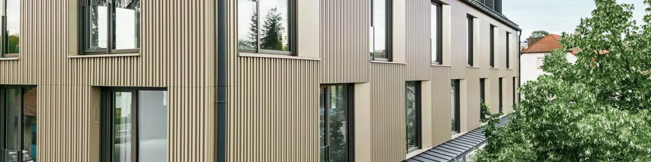 Na obrázku je moderní budova ve Feldkirchu, kterou navrhl architekt Stefan Heigl. Fasáda je vyrobena z Prefalz v barvě bronz a sedlová střecha je pokryta Prefalz v černo-šedé. Velká okna poskytují dostatek světla uvnitř, zatímco prosklená část přízemí nabízí příjemný jídelní kout. Výběr kvalitních materiálů a elegantní design dodávají budově moderní a stylový vzhled.