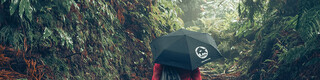 Snímek z lesa s turistkou v červené bundě s deštníkem PREFA a sportovním vakem na zádech symbolizuje ochranu životního prostředí a udržitelnost PREFA, cirkulární ekonomiku a recyklaci