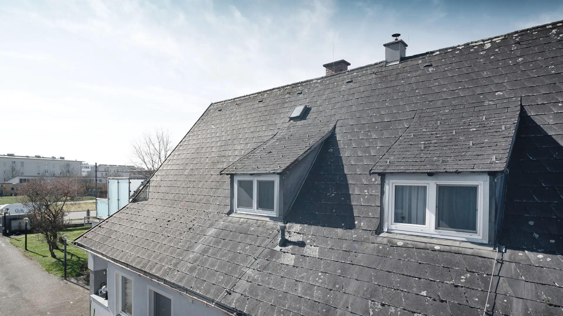 Detailní snímek staré eternitové střechy s viditelnými známkami opotřebení a porostem lišejníků. Střecha má dvě viditelné vikýře. Snímek pořízený před sanací střechy.