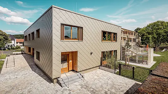 Fotografie ukazuje městskou školku v Alzenau z ptačí perspektivy a zdůrazňuje její moderní architekturu s výraznou hliníkovou fasádou PREFA v bronzové barvě. Fasáda se skládá z kosočtvercových stěnových panelů o rozměrech 44 x 44 cm, které vytvářejí harmonický, šupinovitý vzor a propůjčují budově elegantní dynamiku. Velká dřevěná okna s vertikálními žaluziemi dodávají teplé akcenty a propouštějí dostatek přirozeného světla. Napravo od snímku se otevírá pozemek s oploceným hřištěm, zelenými plochami a krytou terasou. Kombinace odolného, ​​bezúdržbového hliníku a udržitelných materiálů činí z budovy ukázkový projekt funkční a ekologické architektury ve vzdělávací výstavbě.