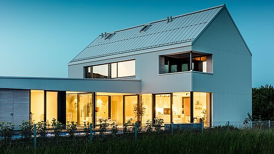 Obrázek ukazuje moderní rodinný dům ve Freistadtu z perspektivy, která zdůrazňuje harmonickou souhru architektury a přírody. Výrazná fasáda PREFALZ v čistě bílé barvě okamžitě upoutá pozornost, zatímco velkorysé prosklené plochy vytvářejí otevřený a příjemný interiér. Strmá sedlová střecha, rovněž pokrytá hliníkem PREFALZ, dodává budově dynamický vzhled a chrání ji před povětrnostními vlivy. Stylový, minimalistický design je zdůrazněn teplými dřevěnými prvky na balkonu a terase, které vytvářejí příjemný kontrast. Budova je nejen esteticky příjemná, ale také funkční a udržitelná díky odolným a nenáročným hliníkovým produktům PREFALZ.