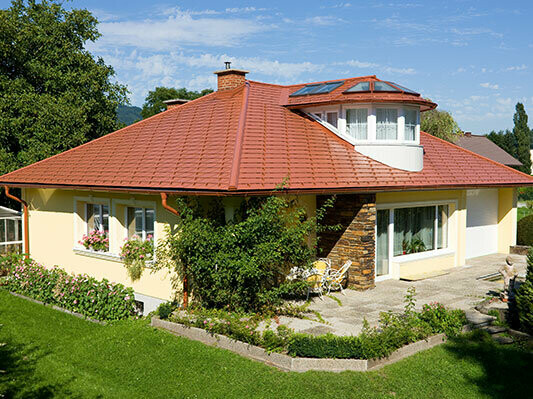 Rodinný dům s valbovou střechou a vikýřem pokrytý PREFA hliníkovými falcovanými šindeli s optikou pálené tašky, v cihlově červené barvě.