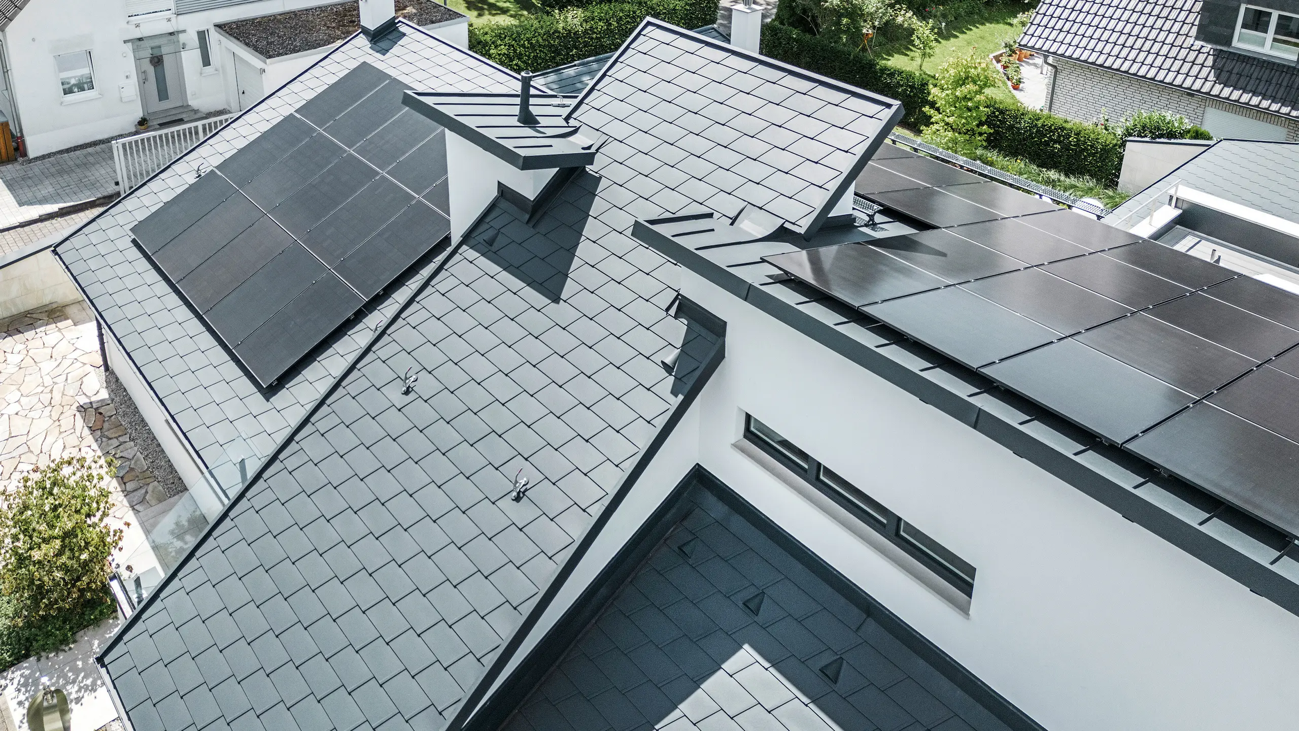 Moderní rodinný dům v Dortmundu s moderním fotovoltaickým systémem instalovaným na střešním systému PREFA, který se skládá ze střešních šindelů DS.19 a krytiny PREFALZ se stojatou drážkou v barvě P.10 antracit. Fotovoltaický systém je namontován pomocí solárních držáků Vario pro střešní šindele a solárních držáků PREFALZ Vario pro plechy se stojatou drážkou. Solární systém zajišťuje efektivní výrobu energie z obnovitelných zdrojů. Propracovaný design střechy nabízí optimální rovnováhu mezi estetikou a funkčností.  Translated with DeepL.com (free version)