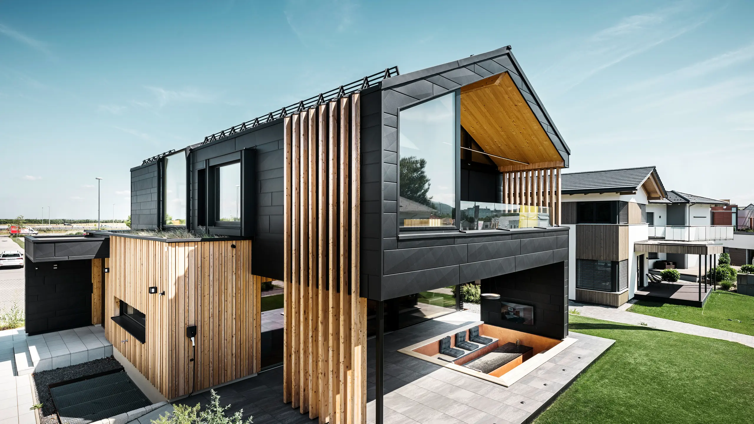 [Translate to Český:] Seitenansicht des Musterhauses Autark in der Blauen Lagune in Vösendorf, Österreich. Das Obergeschoss besticht durch große Fensterflächen und eine moderne Aluminiumfassade aus PREFA Siding.X Paneelen in P.10 Schwarz, kombiniert mit vertikalen Holzelementen. Die großzügige Terrasse im Erdgeschoss mit eingelassener Sitzgelegenheit fügt sich nahtlos in die Architektur ein. Das stilvolle und nachhaltige Design des Hauses verbindet Aluminium und Holz auf eindrucksvolle Art und Weise.