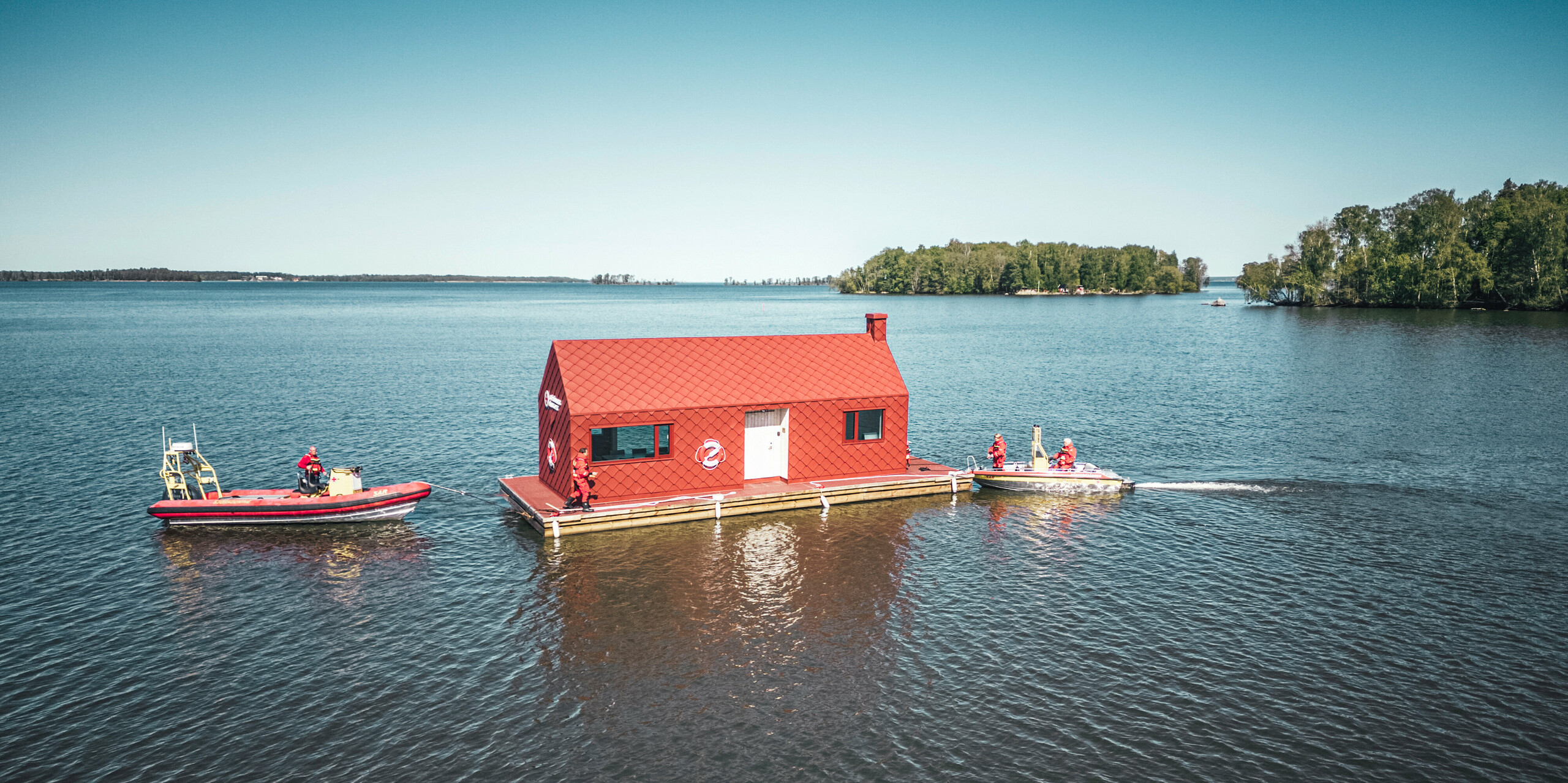 Letecký pohled na dům vodní záchranné stanice na jezeře Hjälmaren ve Švédsku, obklopený klidnou vodní plochou. Nápadná, tmavě červená budova, vybavená PREFA falcovanými a fasádními šablonami 29×29, trůní na plovoucím těle a je připravena nabídnout útočiště a operační základnu. Vedle nádražního domu, který vyniká svou charakteristickou barvou a tvarem, hlídkují dva záchranné čluny, které symbolizují neustálou ostražitost a připravenost vodních záchranářů.