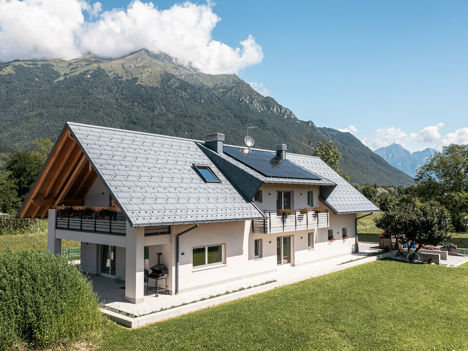 Záběr z dronu na rodinný dům v Belluno, Itálie, pokrytý střešním panelem PREFA R.16 v barvě P.10 antracit. Nemovitost je zasazena do úchvatné alpské krajiny a kombinuje tradiční prvky, jako jsou odhalené dřevěné štíty, s moderními, odolnými stavebními materiály, čímž vzniká dokonalá symbióza tradice a moderny.