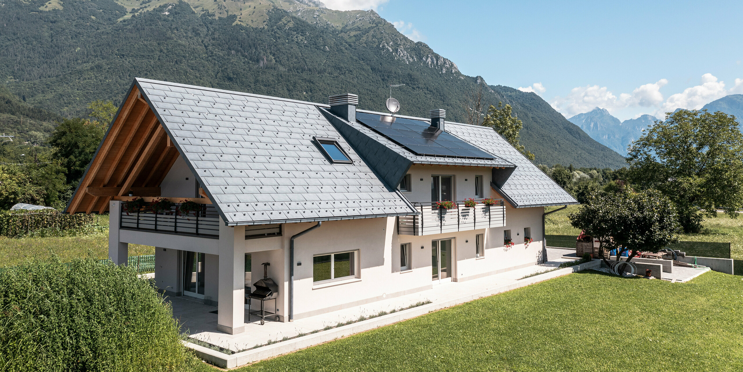 Záběr z dronu na rodinný dům v Belluno, Itálie, pokrytý střešním panelem PREFA R.16 v barvě P.10 antracit. Nemovitost je zasazena do úchvatné alpské krajiny a kombinuje tradiční prvky, jako jsou odhalené dřevěné štíty, s moderními, odolnými stavebními materiály, čímž vzniká dokonalá symbióza tradice a moderny.