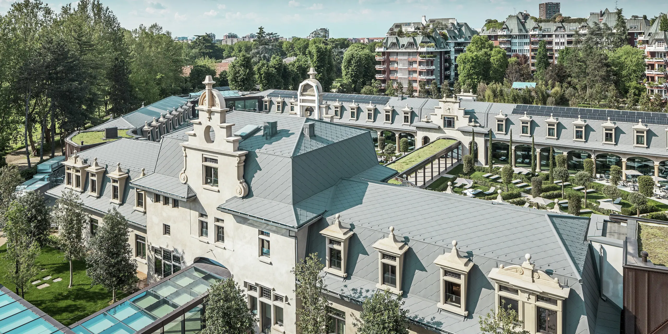 Celkový pohled na impozantní komplex „De Montel – Terme Milano“ s památkově chráněnou secesní architekturou a moderním wellness prostředím. Rozsáhlá střešní krytina z PREFA falcovaných šablon 44 × 44 v barvě P.10 světle šedá dodává komplexu elegantní jednotnost. Integrované fotovoltaické panely, zelené střechy a prosklené spojovací prvky mezi budovami odrážejí požadavky na udržitelnost a moderní design v městském kontextu.