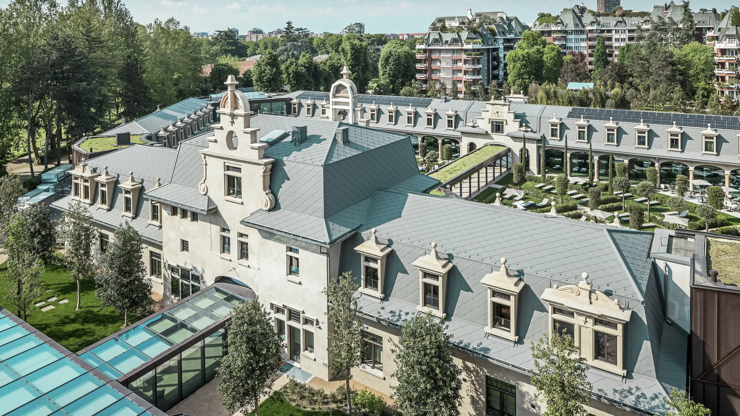 Celkový pohled na impozantní komplex „De Montel – Terme Milano“ s památkově chráněnou secesní architekturou a moderním wellness prostředím. Rozsáhlá střešní krytina z PREFA falcovaných šablon 44 × 44 v barvě P.10 světle šedá dodává komplexu elegantní jednotnost. Integrované fotovoltaické panely, zelené střechy a prosklené spojovací prvky mezi budovami odrážejí požadavky na udržitelnost a moderní design v městském kontextu.