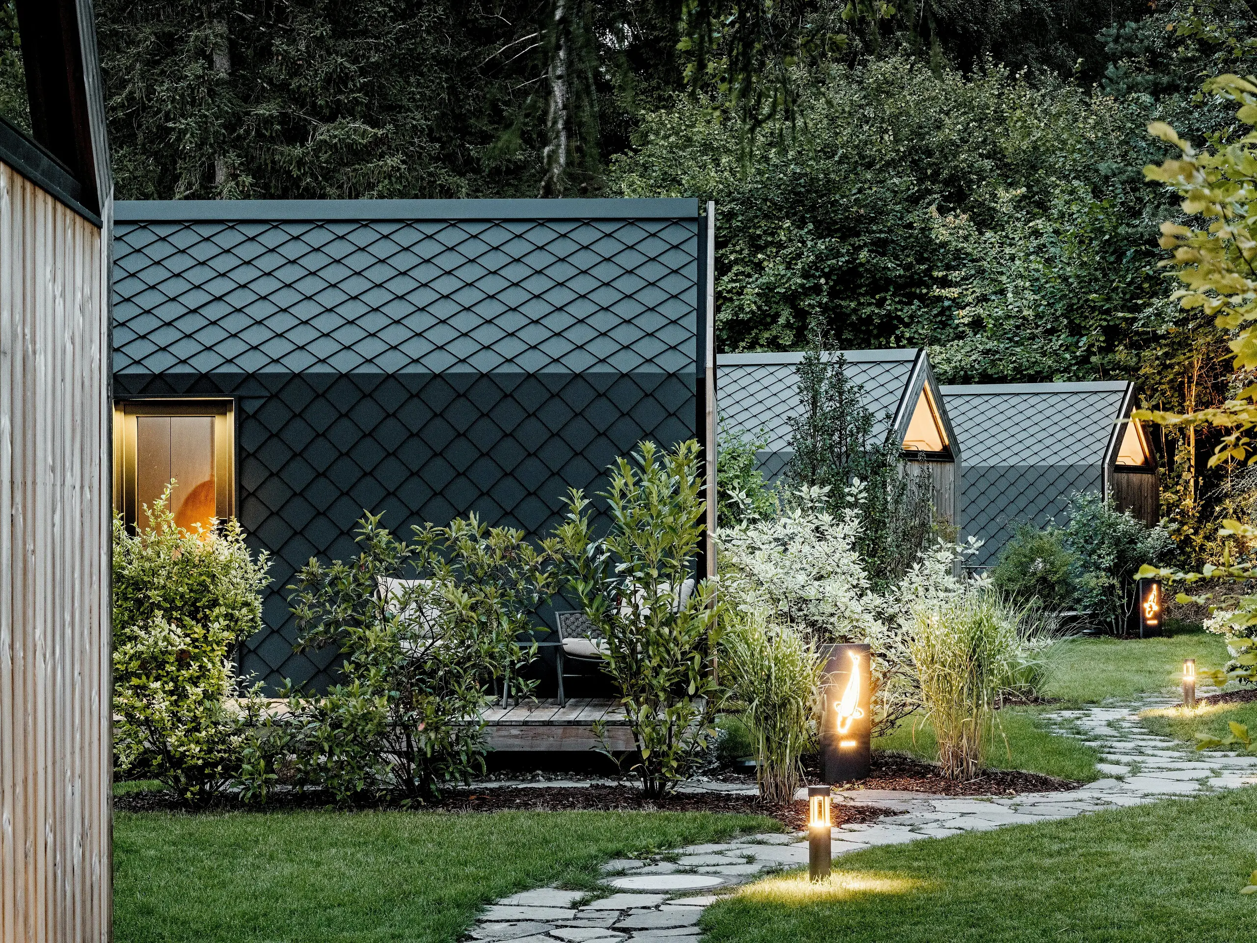 Osvětlená zahradní cesta vede glampingovým resortem Secret Koi Garden u jezera Natterer k několika minimalistickým domkům. Kompaktní konstrukce jsou kompletně obloženy hliníkovými střešními a stěnovými panely PREFA v antracitové barvě P.10, jejichž textura rybích šupin vytváří ve večerním světle elegantní efekt. Uprostřed bujné zeleni spojují odolné a povětrnostním vlivům odolné hliníkové povrchy PREFA vysoce kvalitní architekturu s přírodou, klidem a udržitelným designem.