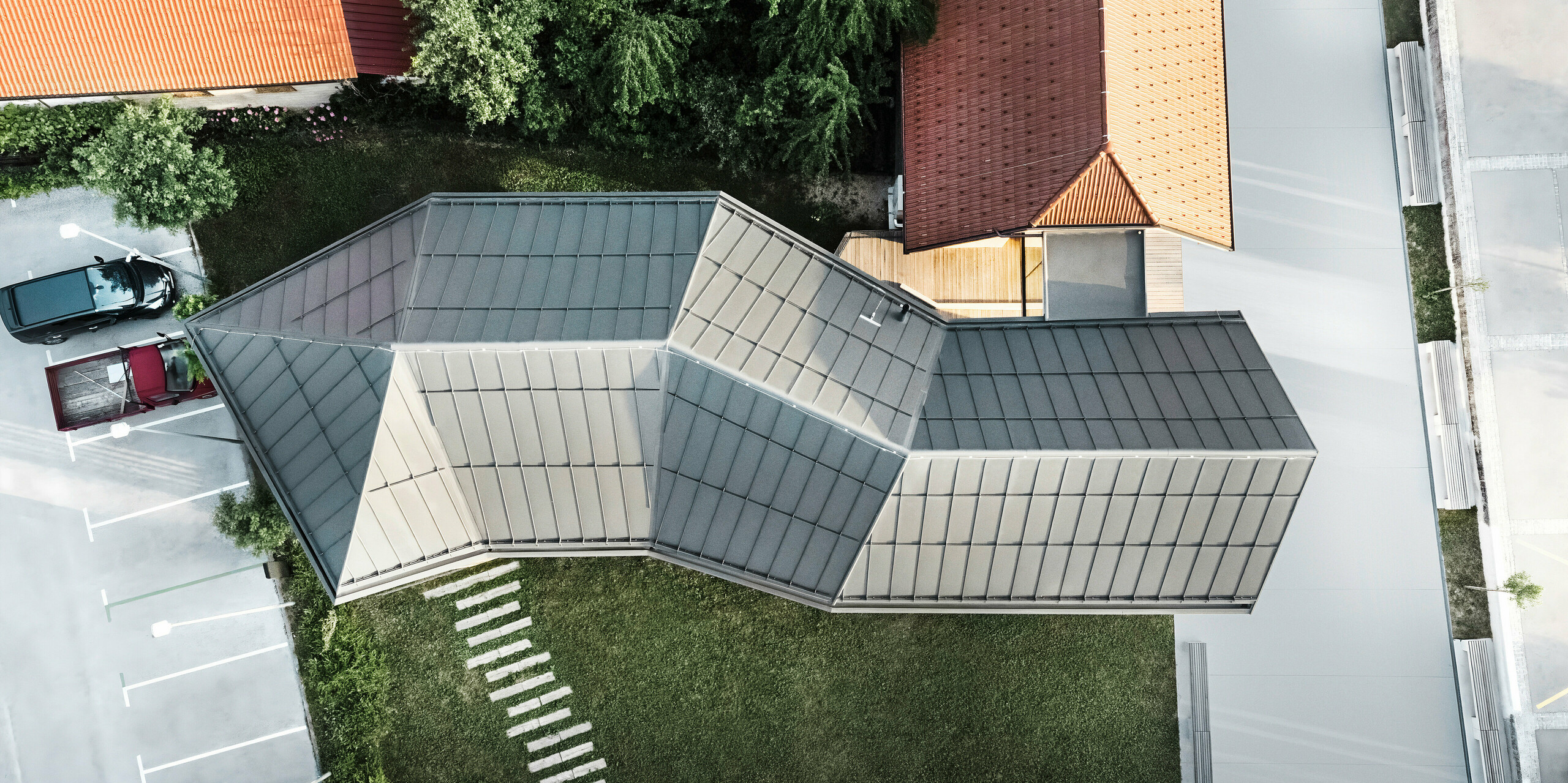 Pohled z ptačí perspektivy na návštěvnické centrum v Ig ve Slovinsku, které zaujme svou složitou geometrickou střechou PREFALZ v P.10 tmavě šedé. Nápadná střešní konstrukce odráží inovativní architekturu a přizpůsobení rozmanitému prostředí s přilehlými tradičními taškovými střechami. Precizní zpracování a moderní design podtrhují odbornost PREFA v moderním designu střech.