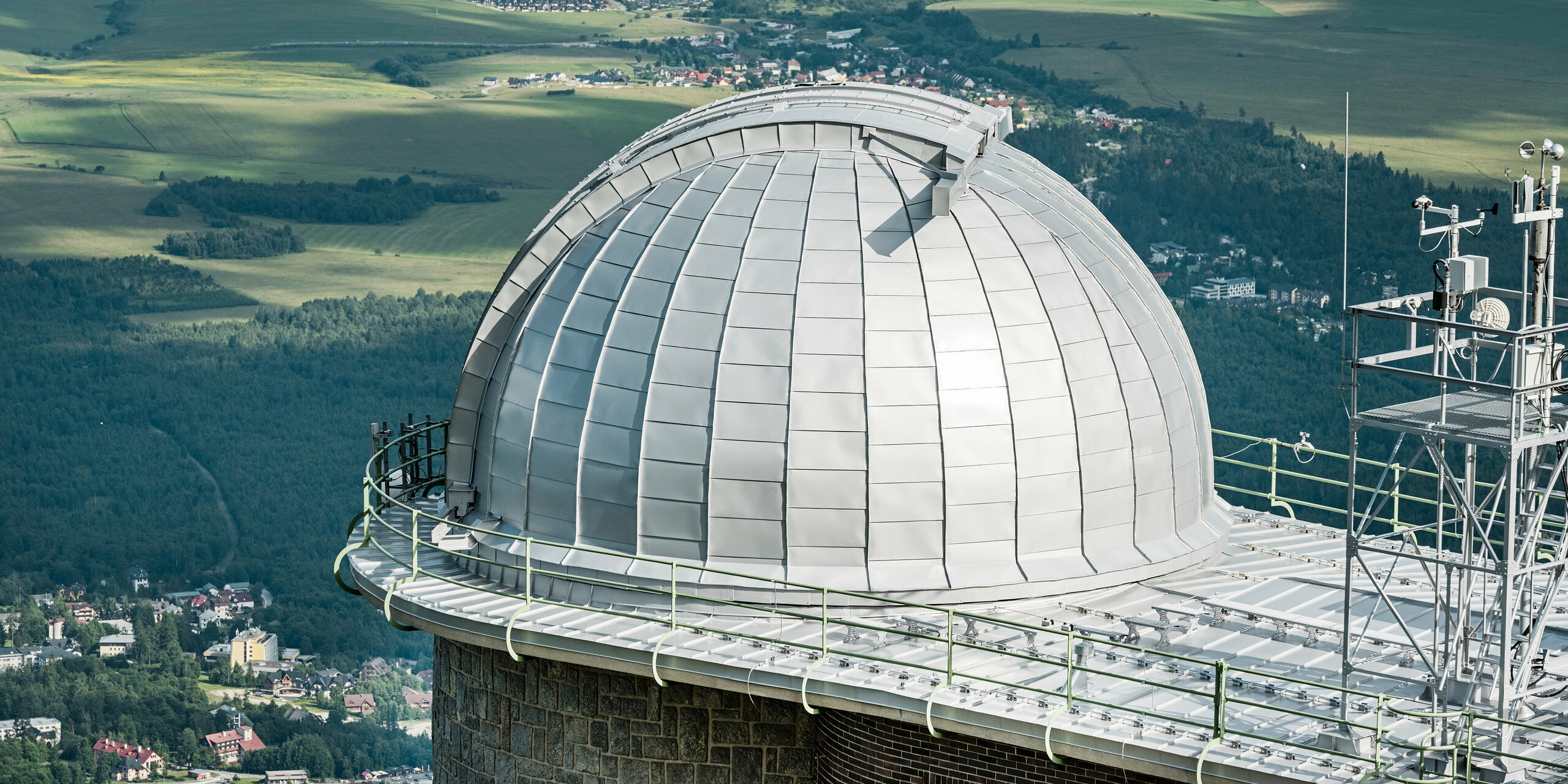 Panoramatický pohled na impozantní kopuli Hvězdárny na Skalnatém Plese, mistrně zasazenou do malebné krajiny Vysokých Tater. Stříbrný metalický hliníkový obklad PREFALZ se třpytí v kontrastu se svěží zelení okolních lesů a jemnými obrysy venkovské krajiny. Tento architektonický skvost, spočívající v nadmořské výšce 1 783 metrů nad mořem, je nejen mezníkem astronomických objevů od roku 1943, ale také svědectvím o odolnosti a ekologické udržitelnosti hliníkových výrobků PREFA, podpořené 40letou zárukou.