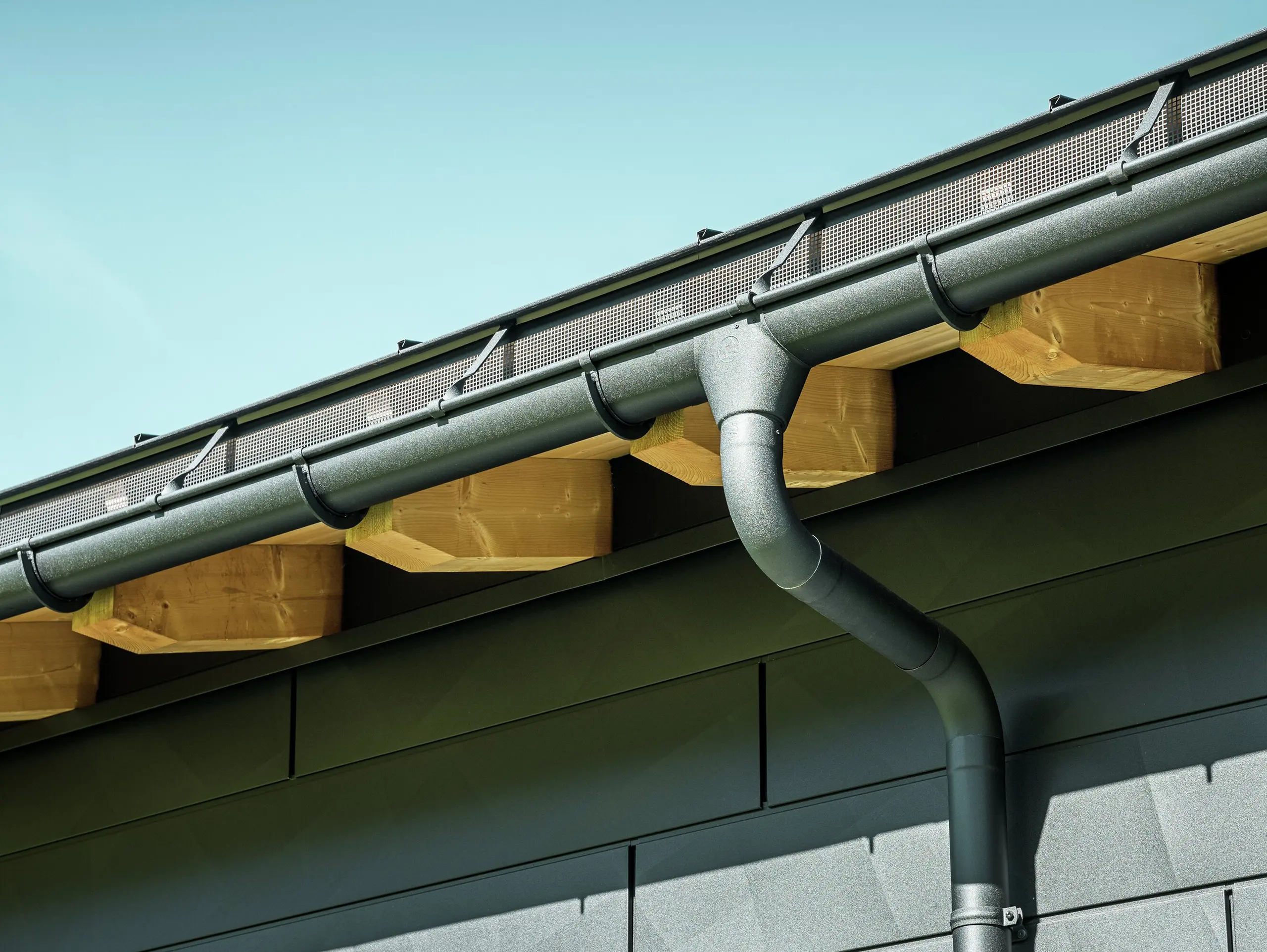 Detailní záběr fasády moderního rodinného domu v Ascharině ve Švýcarsku. Stěny budovy byly obloženy výraznými fasádními panely PREFA Siding.X v barvě P.10 černá. Barevně sladěný je také okapový systém PREFA s půlkulatým žlabem a kruhovým svodem.