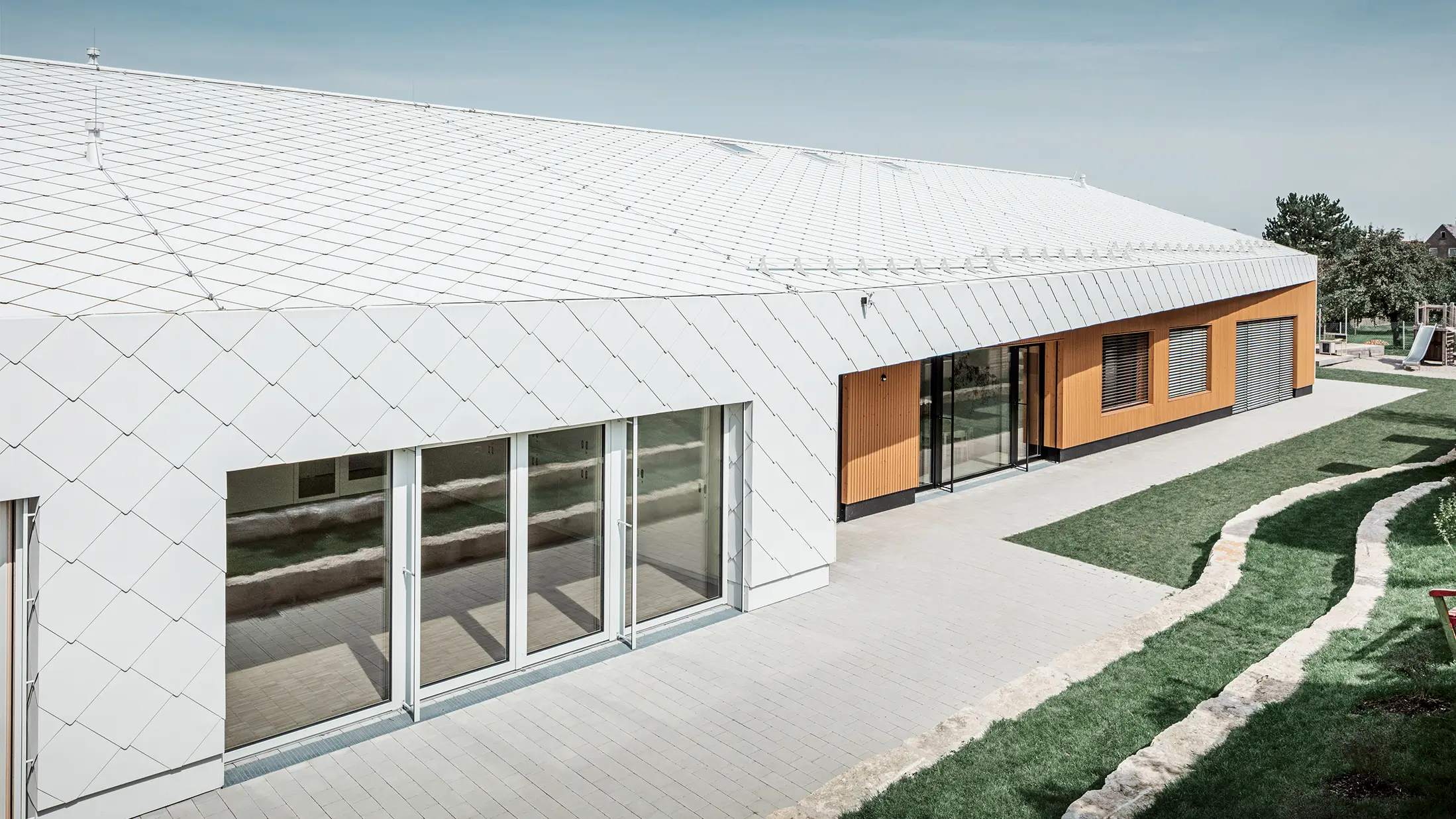Mateřská školka se střechou a fasádními šablonami 44 × 44 v barvě prefa bílá. Střešní plocha pokračuje dále přes fasádu; velká okna