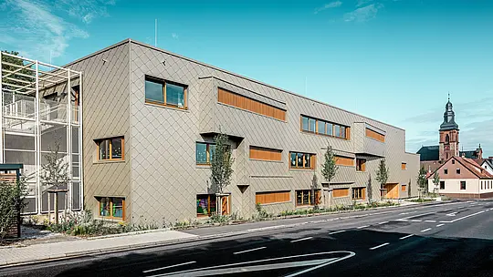 Moderní školka v Alzenau, zalitá zářivým denním světlem, odhaluje v celé své jasnosti svou výraznou hliníkovou fasádu PREFA, složenou z bronzových kosočtverců stěn o rozměrech 44 x 44 cm. Jednotná, šupinovitá struktura, prokládaná stupňovitě uspořádanými okny a vertikálními dřevěnými žaluziemi, dodává fasádě hloubku a rytmus. Vlevo od obrazu je vidět jemné schodiště s kovovou mřížkovou strukturou, které přispívá k pocitu transparentnosti a lehkosti. V pozadí stojí pozdně gotický kostel v Alzenau – symbol harmonického spojení historických stavebních tradic a současné architektury. Bezúdržbový a odolný fasádní systém PREFA podporuje udržitelný, funkční a esteticky sofistikovaný design této progresivní vzdělávací budovy.