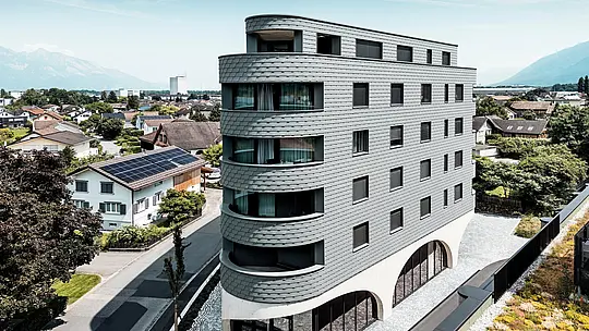 Letecký pohled na Bernerhaus v Buchsu s jeho výrazným trojúhelníkovým tvarem a jemně zaoblenými balkony. Fasáda, obložená fasádními šindeli PREFA v barvě P.10 světle šedé, obaluje budovu jako šupinovitý hliníkový plášť a vytváří živou hru světla a stínu. Nad masivním betonovým základem s elegantními arkádami působí lehká, povětrnostním vlivům odolná hliníková fasáda PREFA mimořádně precizně, trvanlivě a nenáročně na údržbu – moderní poutač uprostřed alpské městské krajiny.