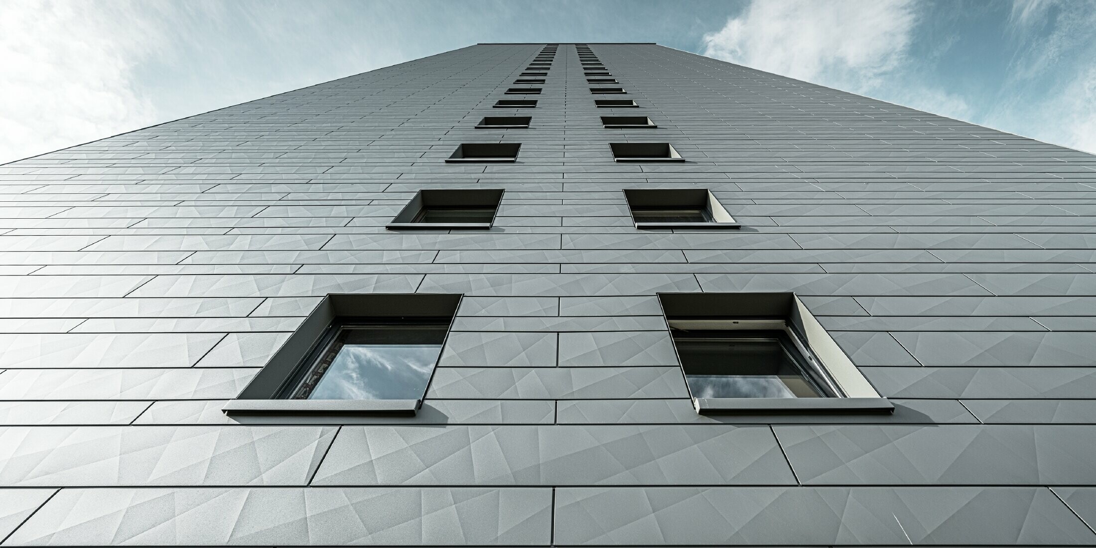 Pohled nahoru na fasádu vícepodlažní výškové budovy, fasáda je obložena horizontálními fasádními lamelami Siding.X od PREFA v barvě P.10 světle šedé