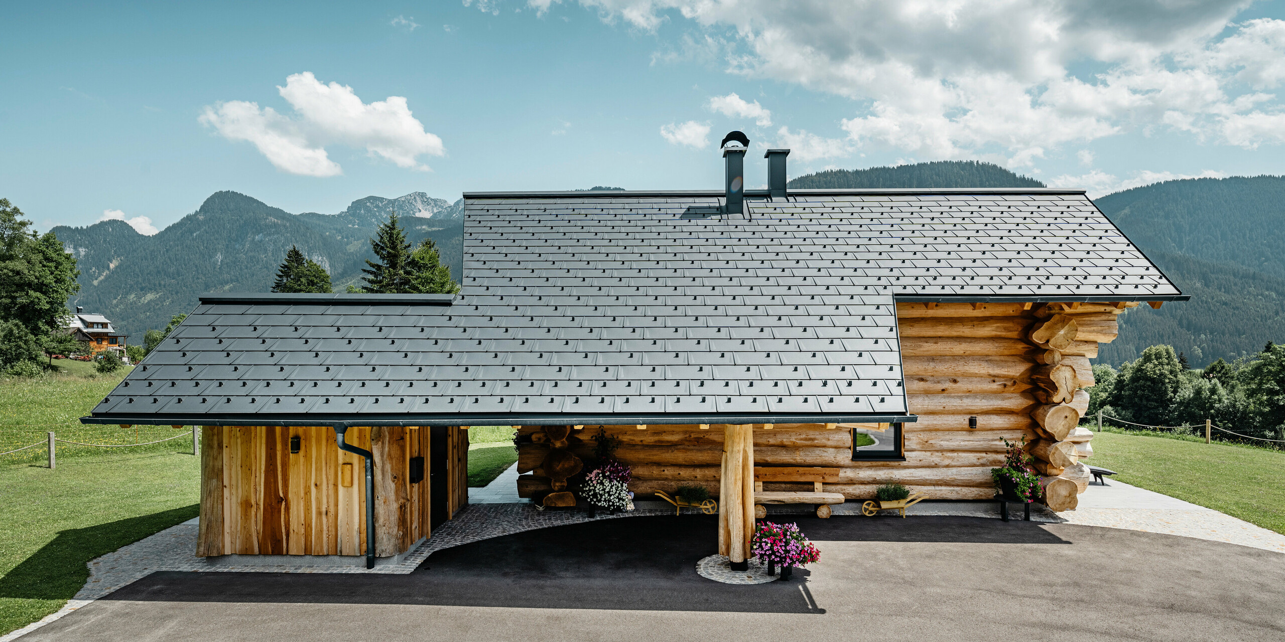 Boční pohled na příjemný srub v rakouském Gosau, vyznačující se robustní hliníkovou střechou PREFA se střešními panely R.16 v barvě P.10 antracitové. Na obrázku je harmonické spojení tradiční blokové výstavby s moderními, vysoce kvalitními střešními krytinami, které odráží dlouhou životnost a estetický design hliníkových výrobků PREFA. Dřevěný dům stojí na pozadí zvlněných zelených kopců a majestátního horského panoramatu, které podtrhuje přírodní okolí.
