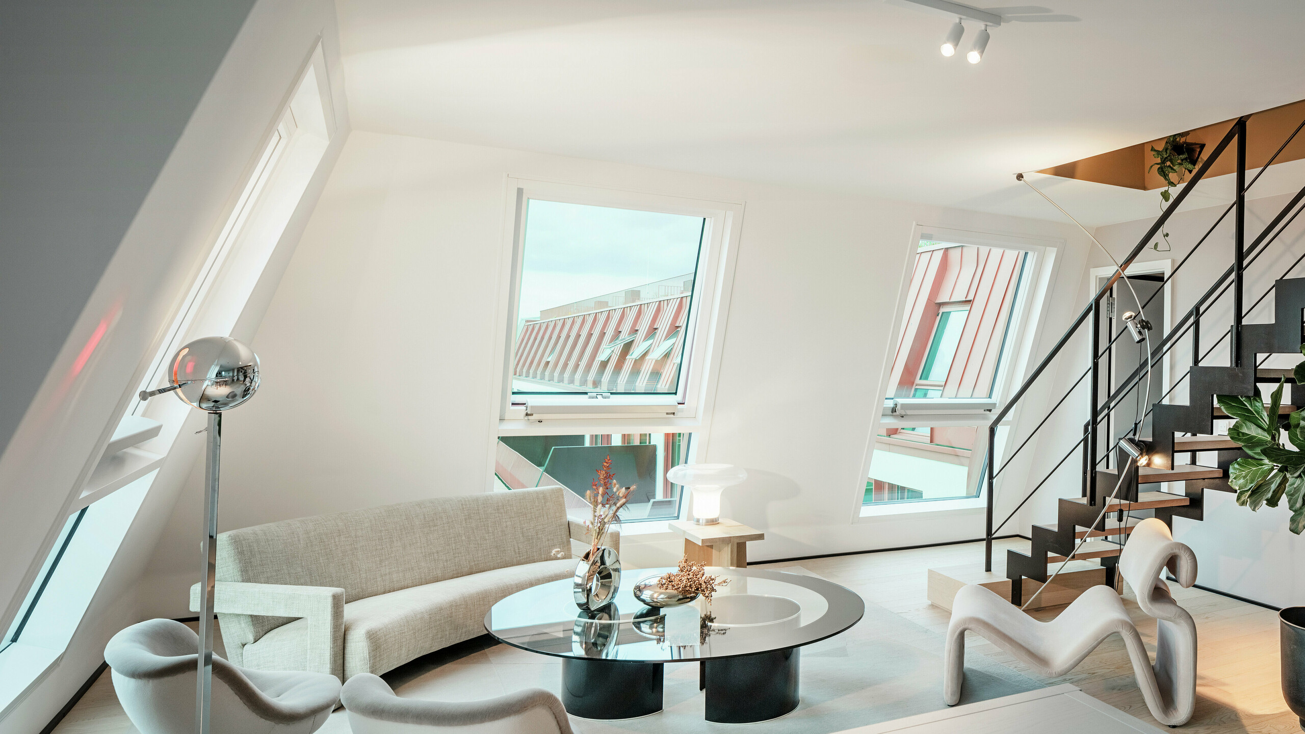 Pohled do interiéru prosvětleného penthousového bytu v rezidenčním projektu Van B v Mnichově s výhledem na vysoce kvalitní hliníkovou střechu PREFALZ ve speciální barvě IGP Orange HDP. Velká okna propouštějí dostatek denního světla a podtrhují moderní koncept bydlení. Jasné linie, designový nábytek a precizní zpracování střešních napojení ukazují, jak lze kvalitní a stylovou architekturu realizovat nejen venku, ale i uvnitř.