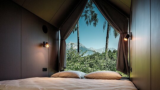 Výhled z postele minimalistického domku v Tajné zahradě Koi u jezera Natterer: Velká trojúhelníková prosklená fasáda otevírá místnost do přírody se stromy a výhledem na tyrolské hory. Jednoduchý interiér přitahuje pozornost ven a podtrhuje koncept zpomalení. Architekturu chrání odolné hliníkové kosočtverce PREFA, které kombinují design, odolnost vůči povětrnostním vlivům a udržitelnost.