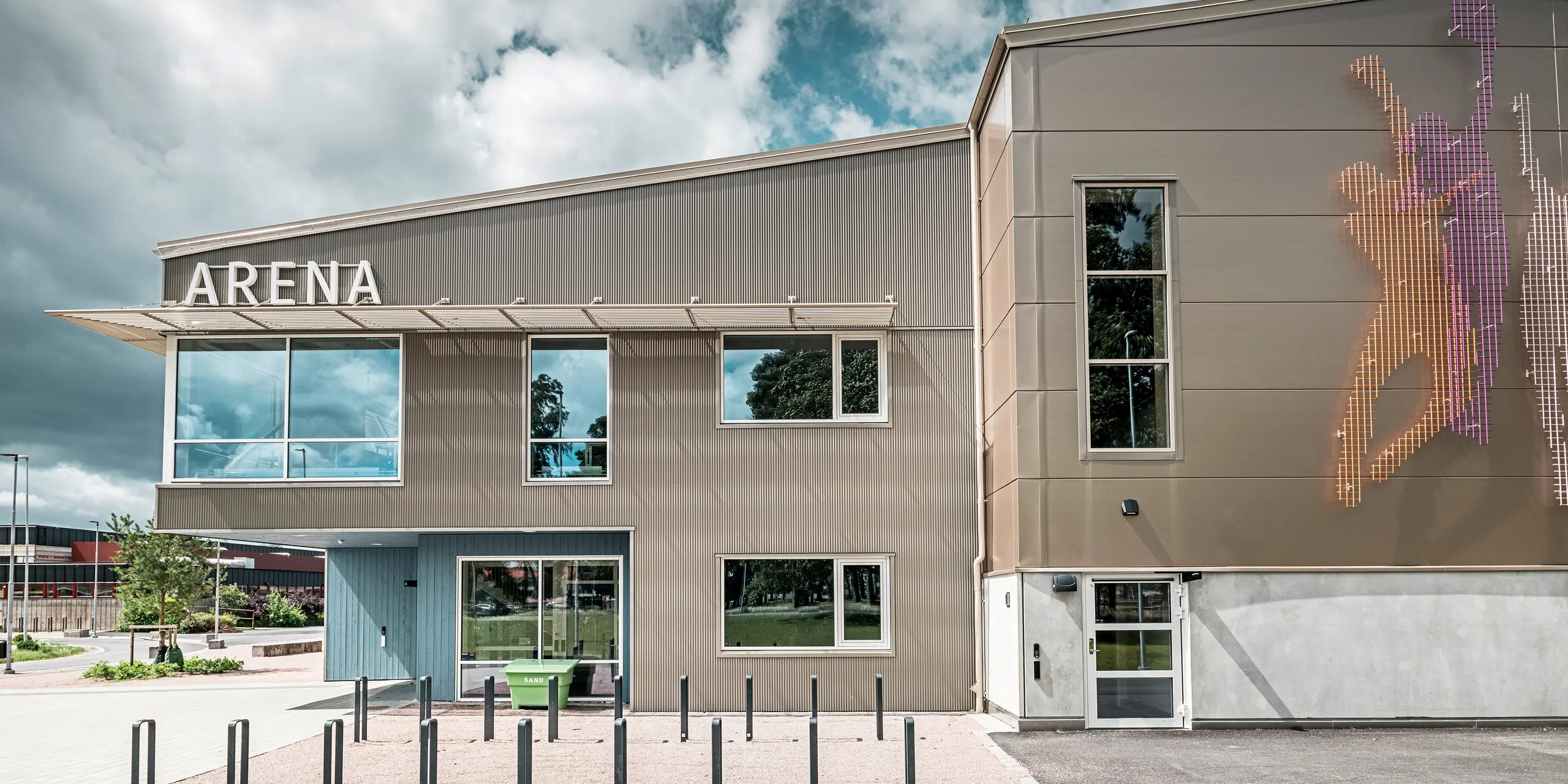 Boční pohled na vstupní prostor do Park Areny ve Värnamo, Švédsko, s moderní fasádou PREFA z pilového Zackenprofilu v bronzu. Hliníková fasáda je odolná a vytváří výrazné světelné a stínové efekty. Vpravo na obrázku je umělecky řešená nástěnná malba, která zobrazuje sportovní postavy a podtrhuje dynamickou atmosféru sportovní haly. Velké okenní plochy propouštějí do budovy dostatek světla a zdůrazňují současný design.