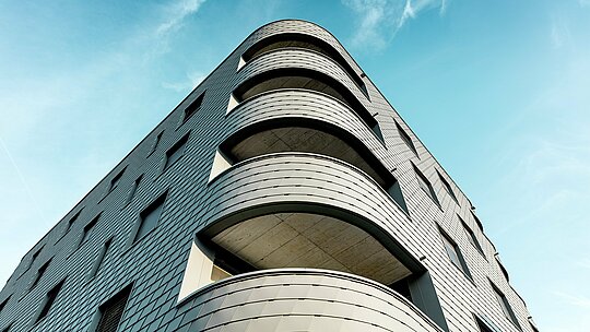 Pohled na spodní stranu moderní bytové a komerční budovy s výrazným zaobleným rohem a splývavými balkony. Šupinatá fasáda z PREFA falcovaných šindelů v barvě P.10 světle šedé se překrývá s budovou jako precizně vyrobený hliníkový plášť a zdůrazňuje jemné křivky. Homogenní povrch podtrhuje čisté linie, zatímco lehký a povětrnostním vlivům odolný hliník PREFA zaručuje maximální rozměrovou stabilitu, trvanlivost a nízkou údržbu i u ostrých křivek – architektonický poutač na pozadí jasně modré oblohy.