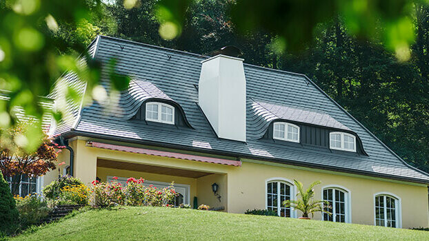 Rodinný dům s čerstvě sanovanou střechou za použití PREFA falcovaných střešních šindelů antracitové barvy, s obloukovými vikýři a bílým komínem.