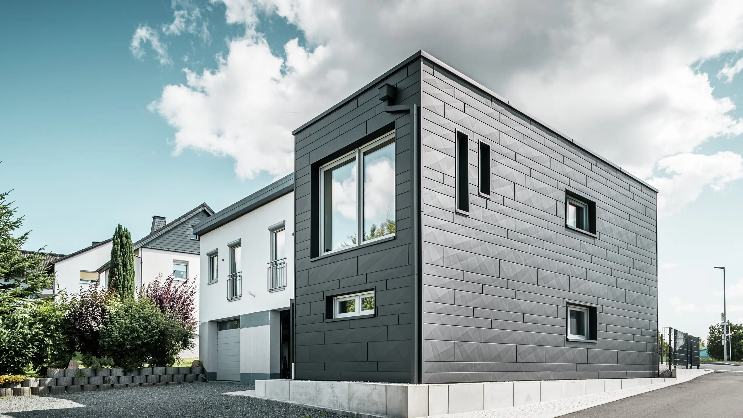 Stylová a moderní fasáda zrekonstruovaného rodinného domu v Teuschnitzu je tvořena jasně strukturovanými panely PREFA Siding.X v barvě P.10 černá. Geometrické uspořádání vysoce kvalitních plechových panelů dodává budově výraznou, nadčasovou estetiku. Okna jsou orámována černými hliníkovými kompozitními deskami, které harmonicky dotvářejí design. Integrované odvodnění střechy PREFA s hranatým svodem představuje funkční a estetické řešení.