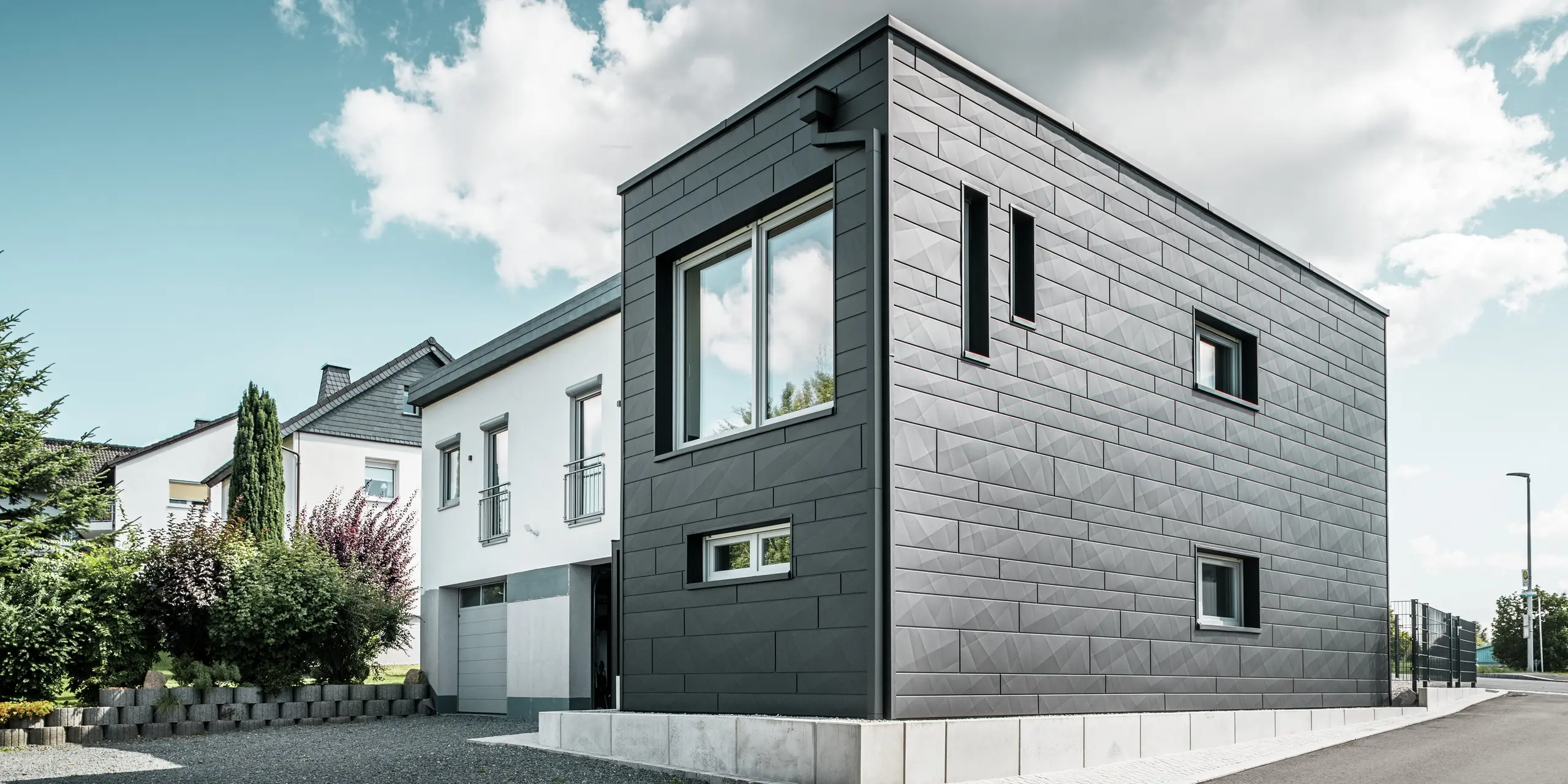 Stylová a moderní fasáda zrekonstruovaného rodinného domu v Teuschnitzu je tvořena jasně strukturovanými panely PREFA Siding.X v barvě P.10 černá. Geometrické uspořádání vysoce kvalitních plechových panelů dodává budově výraznou, nadčasovou estetiku. Okna jsou orámována černými hliníkovými kompozitními deskami, které harmonicky dotvářejí design. Integrované odvodnění střechy PREFA s hranatým svodem představuje funkční a estetické řešení.