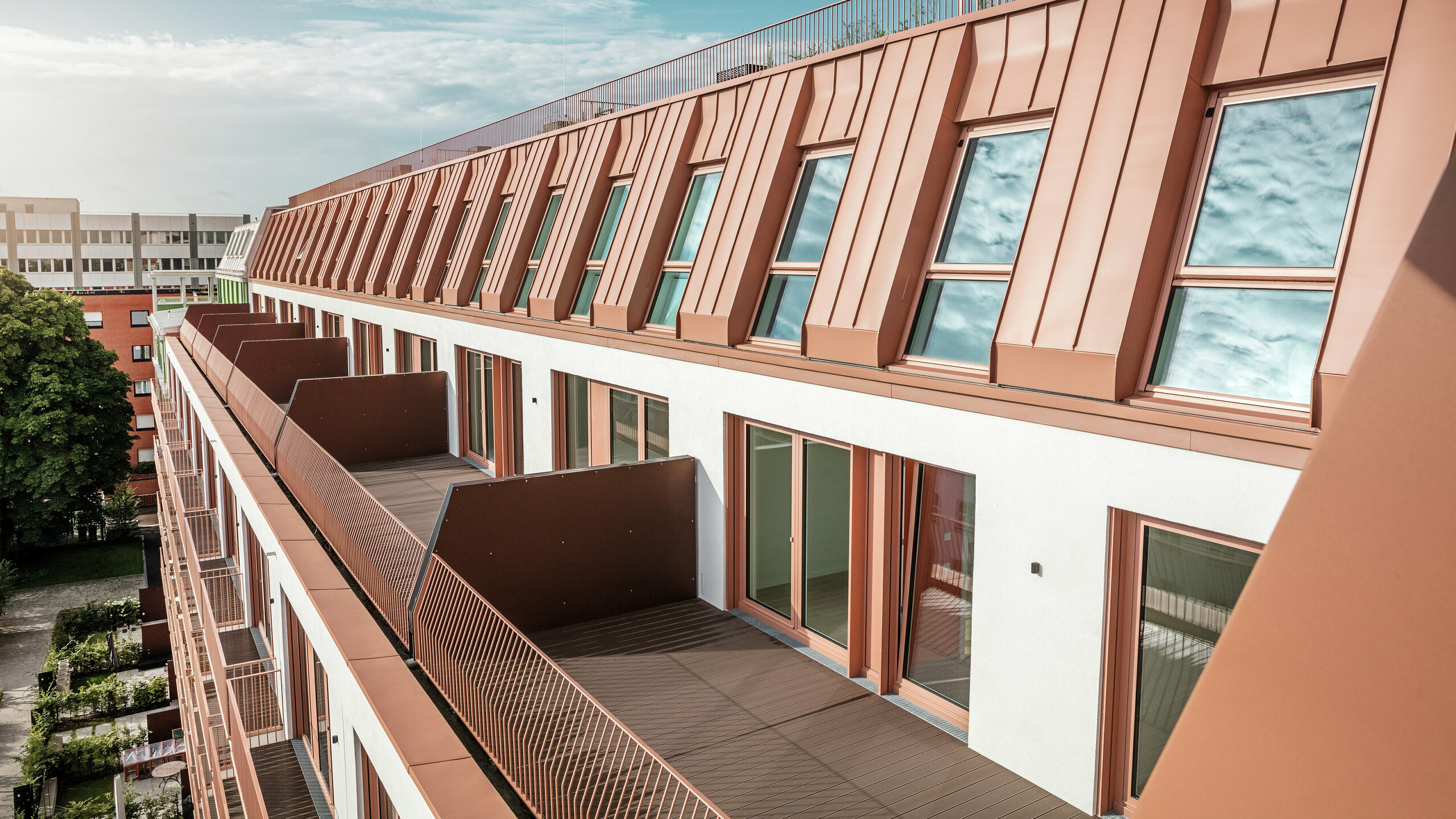 Detailní pohled na moderní design střechy a fasády rezidenčního projektu Van B v Mnichově s vysoce kvalitní hliníkovou střechou PREFALZ z IGP Orange HDP od společnosti PREFA. Precizní zpracování odolného a nenáročného materiálu nabízí optimální ochranu před povětrnostními vlivy a dodává budově jednotný a elegantní vzhled. Velkorá okna a balkony poskytují dostatek přirozeného světla a vytvářejí otevřené, obyvatelné obytné prostředí s architektonickou propracovaností.
