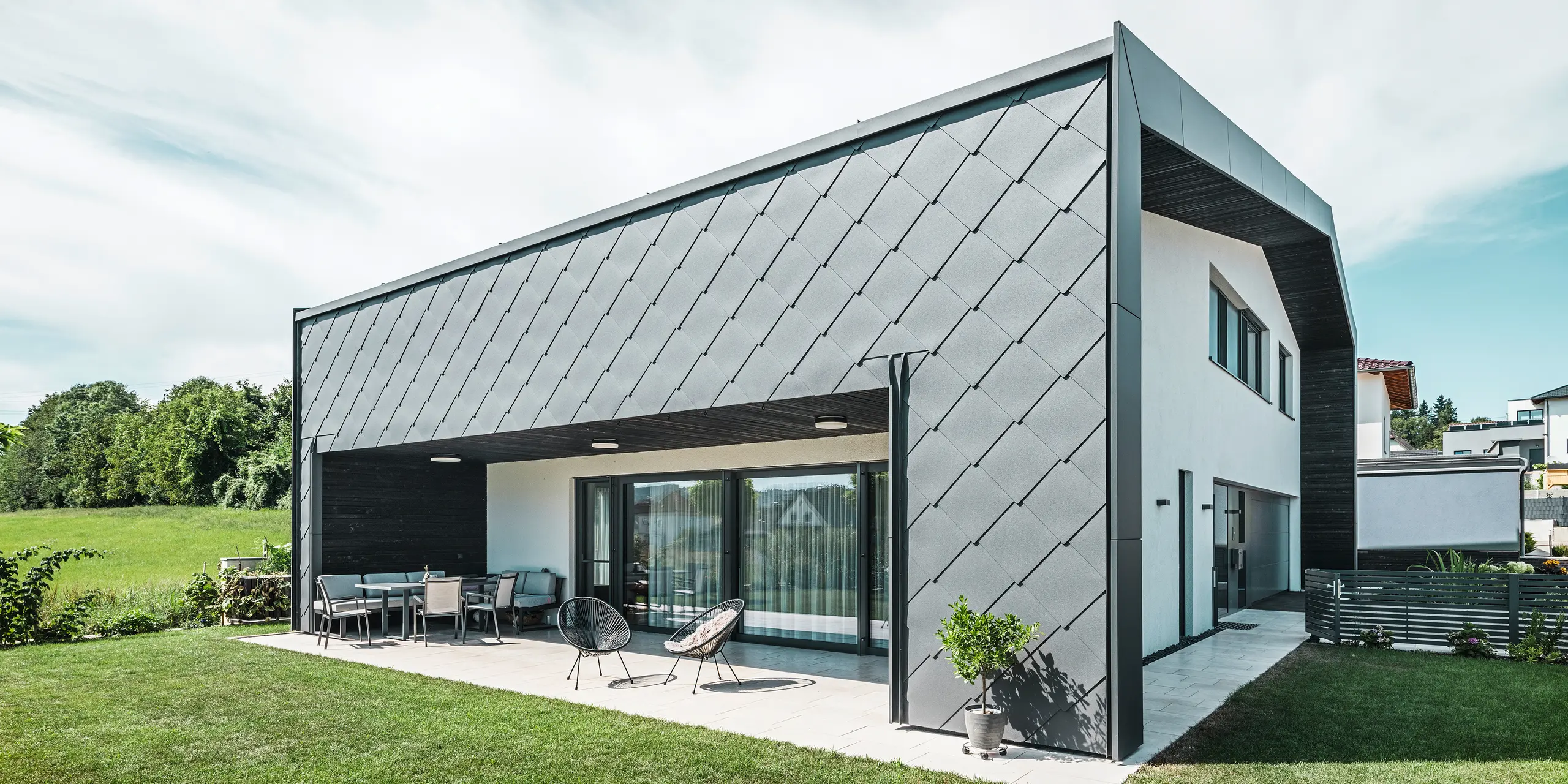 Upravená zahrada rodinného domu v Aschachu. Budova je stylově pokryta tmavě šedými fasádními šablonami PREFA Aluminium 44 × 44. Krytá terasa nabízí příjemné posezení s výhledem na svěží zelený trávník. Elegantní hliníkový obklad v kombinaci s bílými omítkami zajišťuje moderní a povětrnostním vlivům odolnou architekturu. Velké okna propouštějí do interiéru dostatek denního světla a vytvářejí otevřenou, světlou atmosféru.